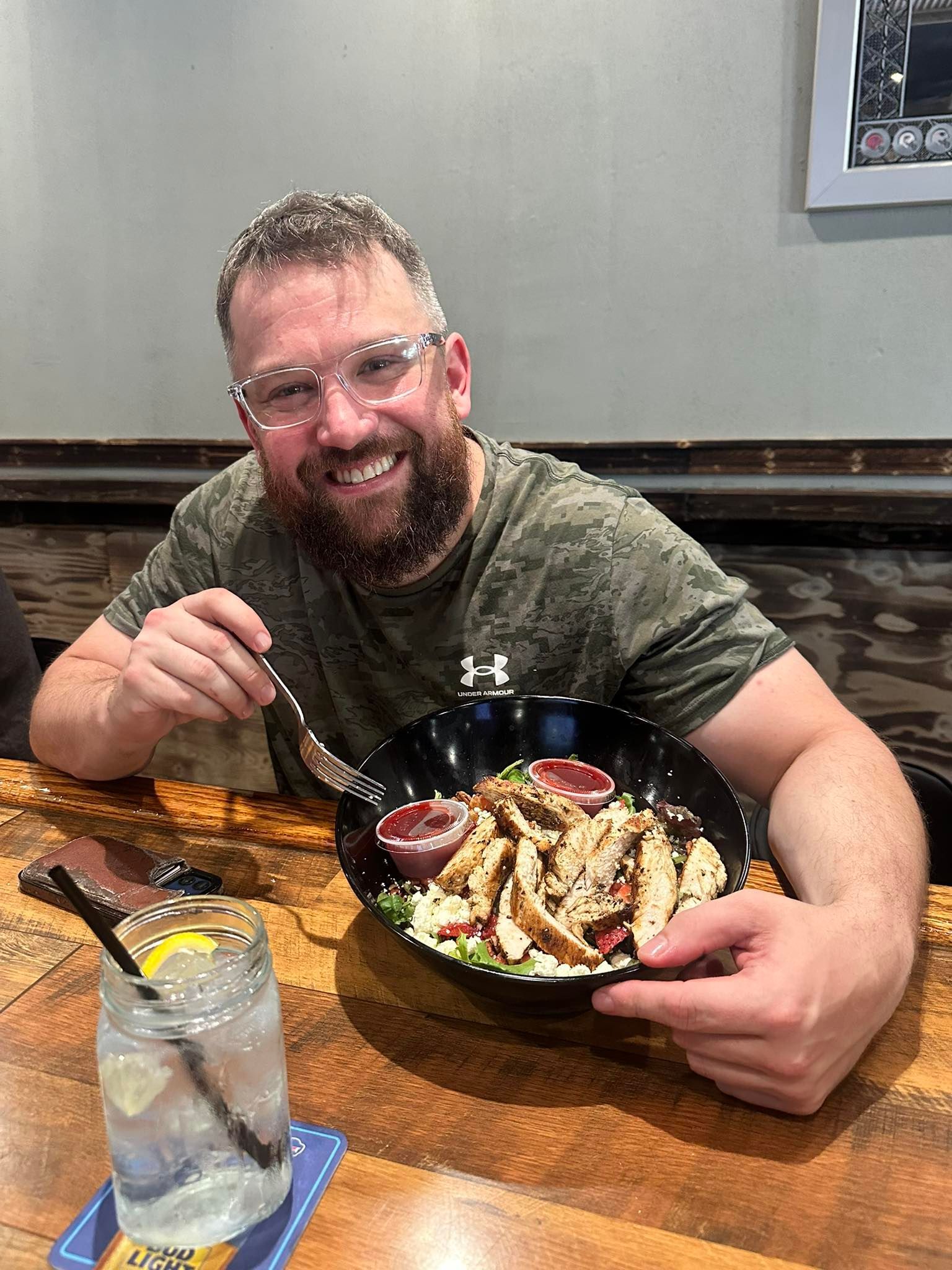 Man with glasses smiles, holding a salad bowl. A drink sits to his left on a wooden table.