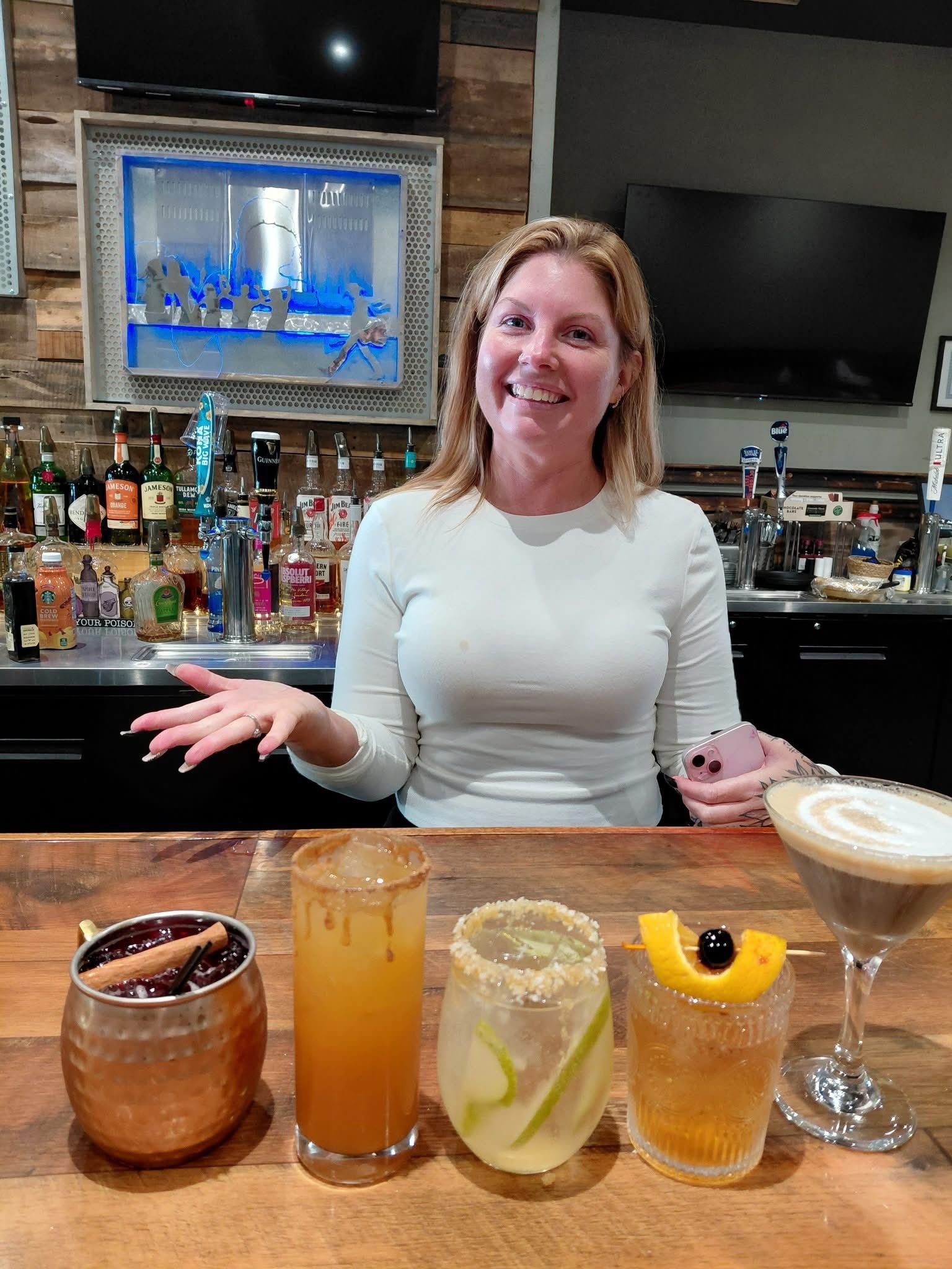 Woman bartender behind bar, smiling, with five cocktails in front.