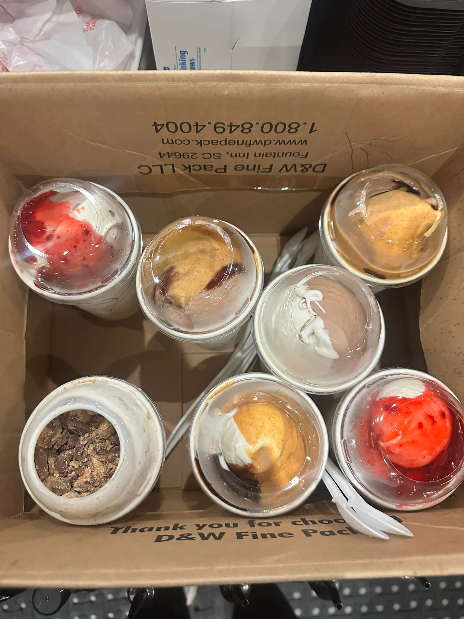 Cardboard box containing seven individual desserts in clear plastic containers with spoons.
