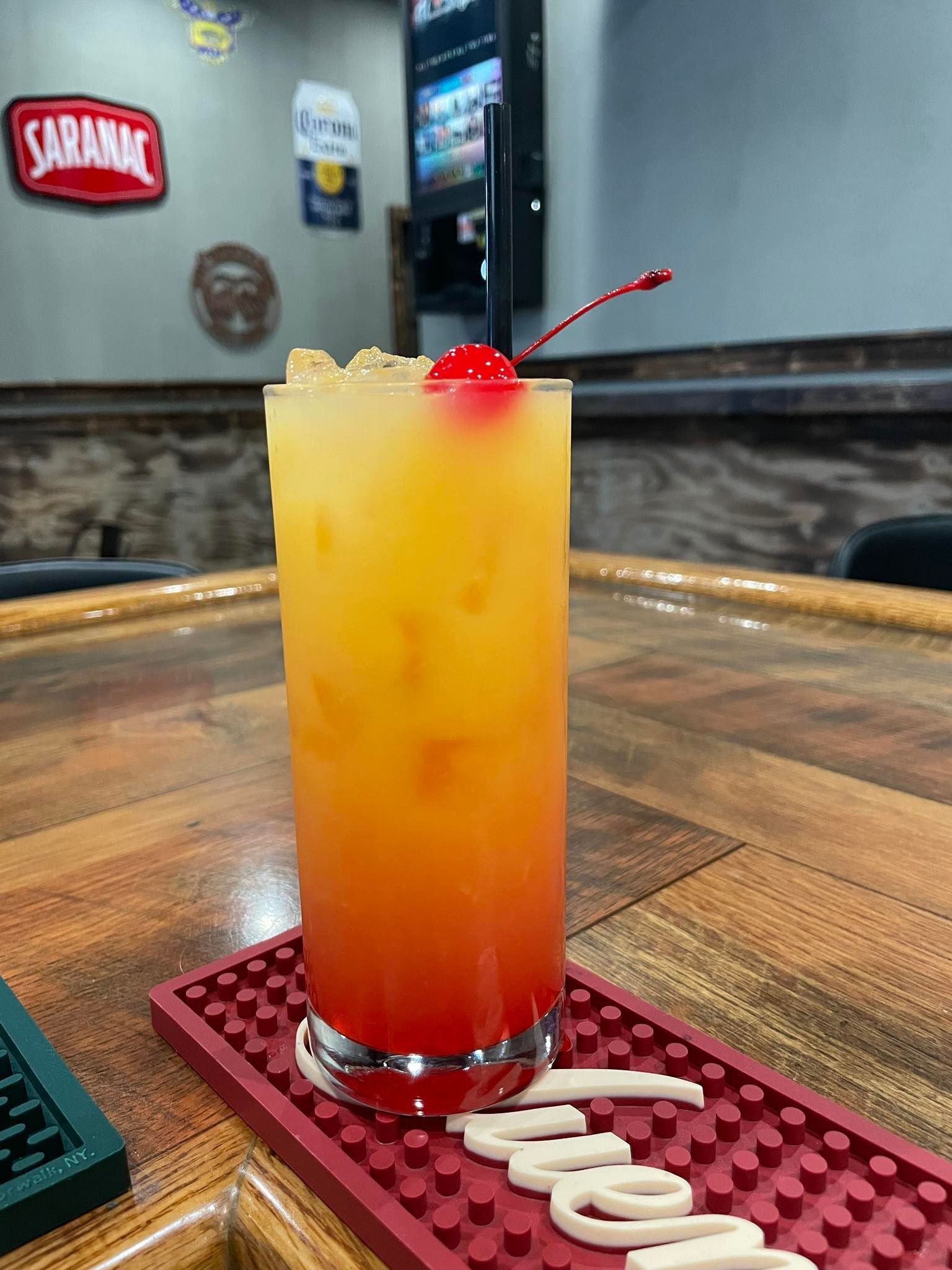 Tall glass with a layered orange cocktail, topped with a cherry, on a bar.