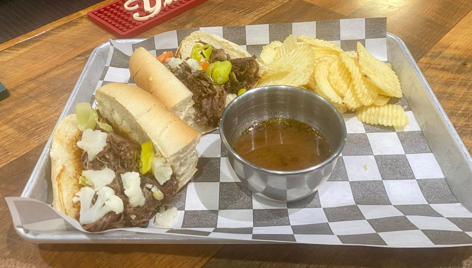 French dip sandwich with chips and au jus on a tray with checkered paper.