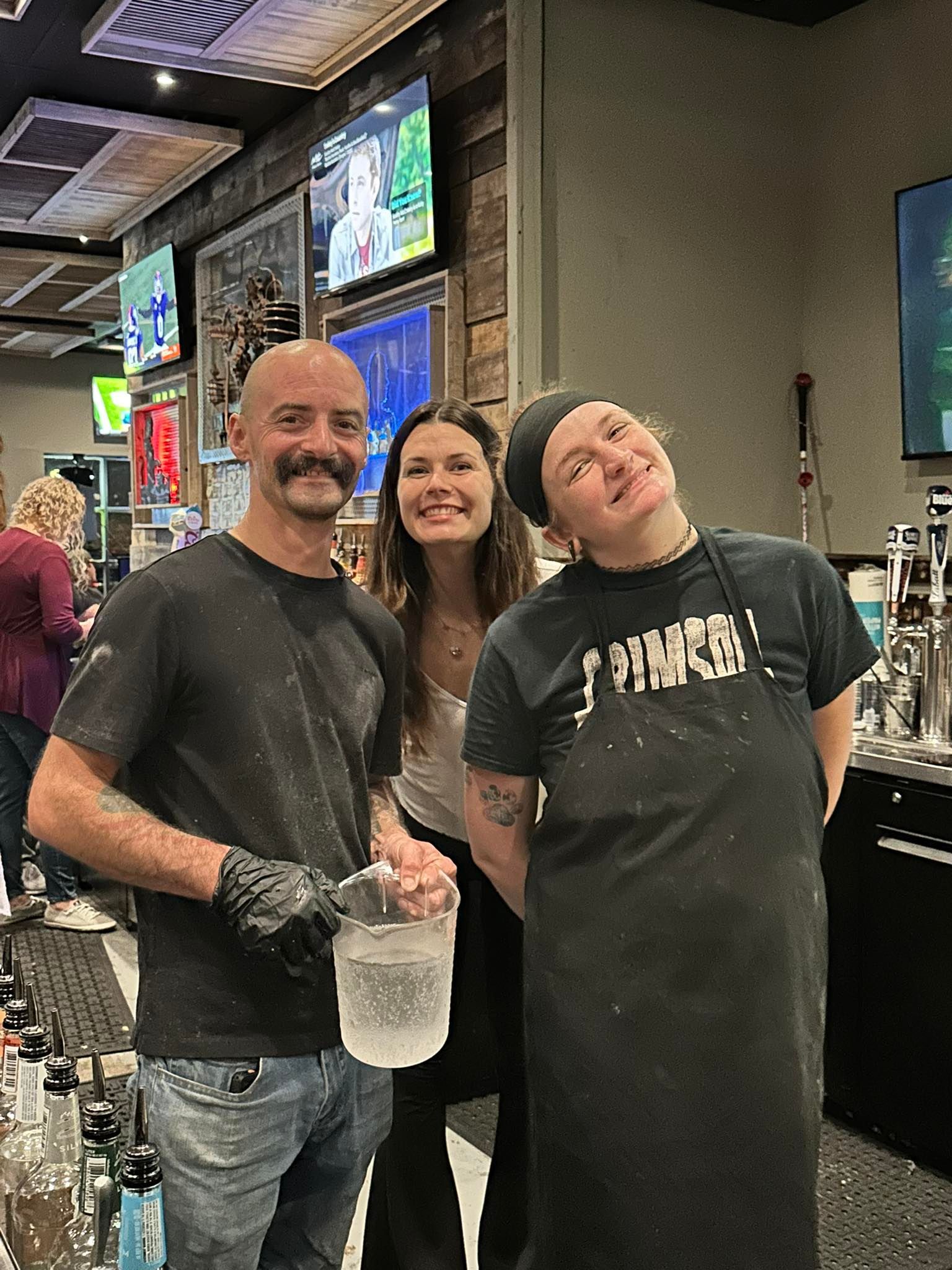 Three people smiling behind a bar. One holds ice, another leans in, and the third wears an apron.