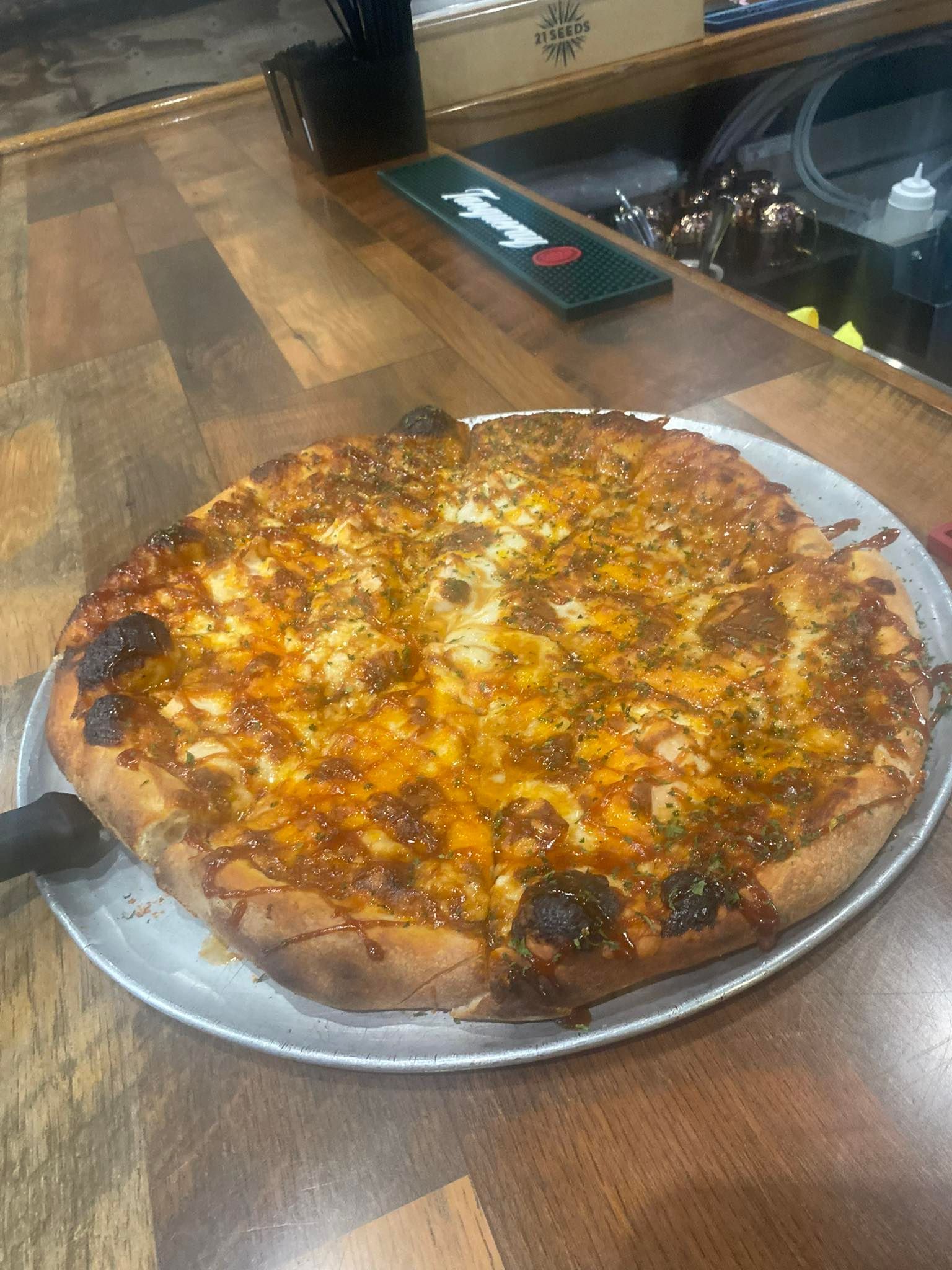A large pizza on a metal tray, sitting on a wooden bar. It is golden brown with toppings.