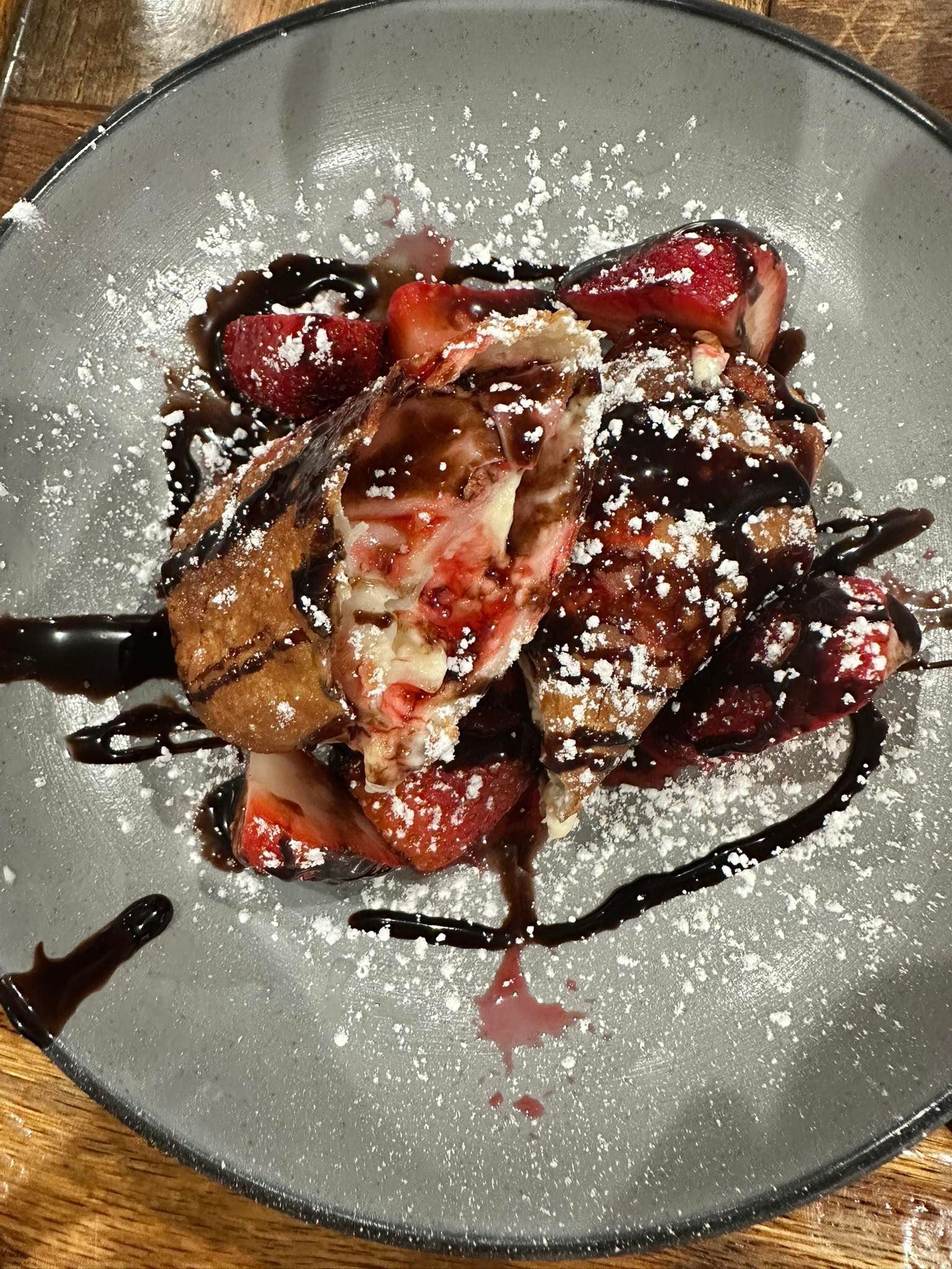Fried pastry with strawberries, chocolate syrup, and powdered sugar on a gray plate.