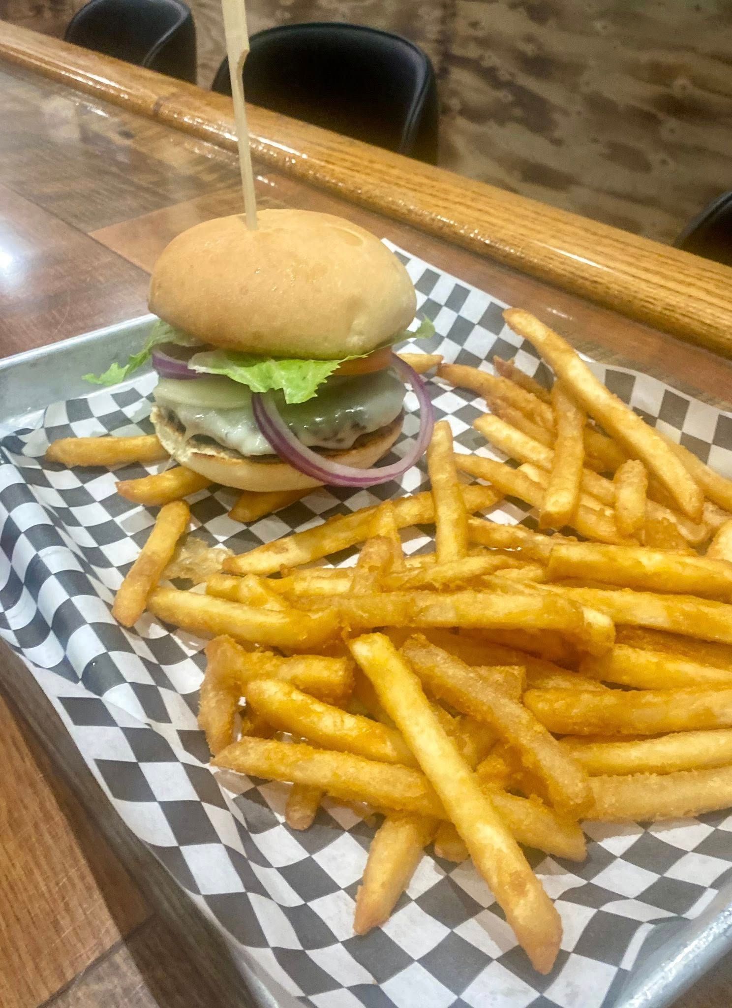 Burger with fries on a checkered paper-lined tray. Wooden skewer secures bun. Fries are golden.