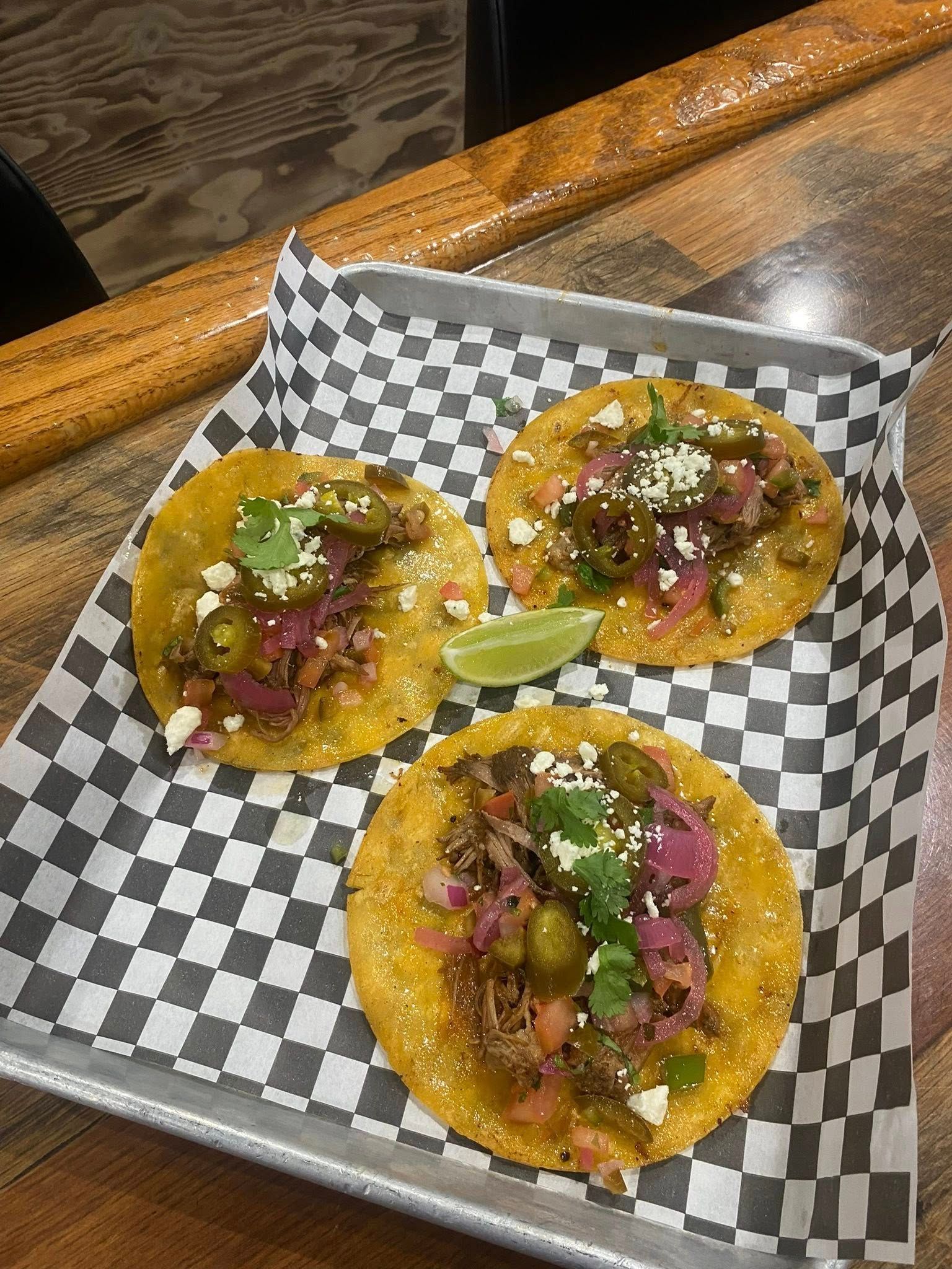 Three tacos on a checkered paper-lined tray. Toppings include cilantro and pickled onions, with a lime wedge.