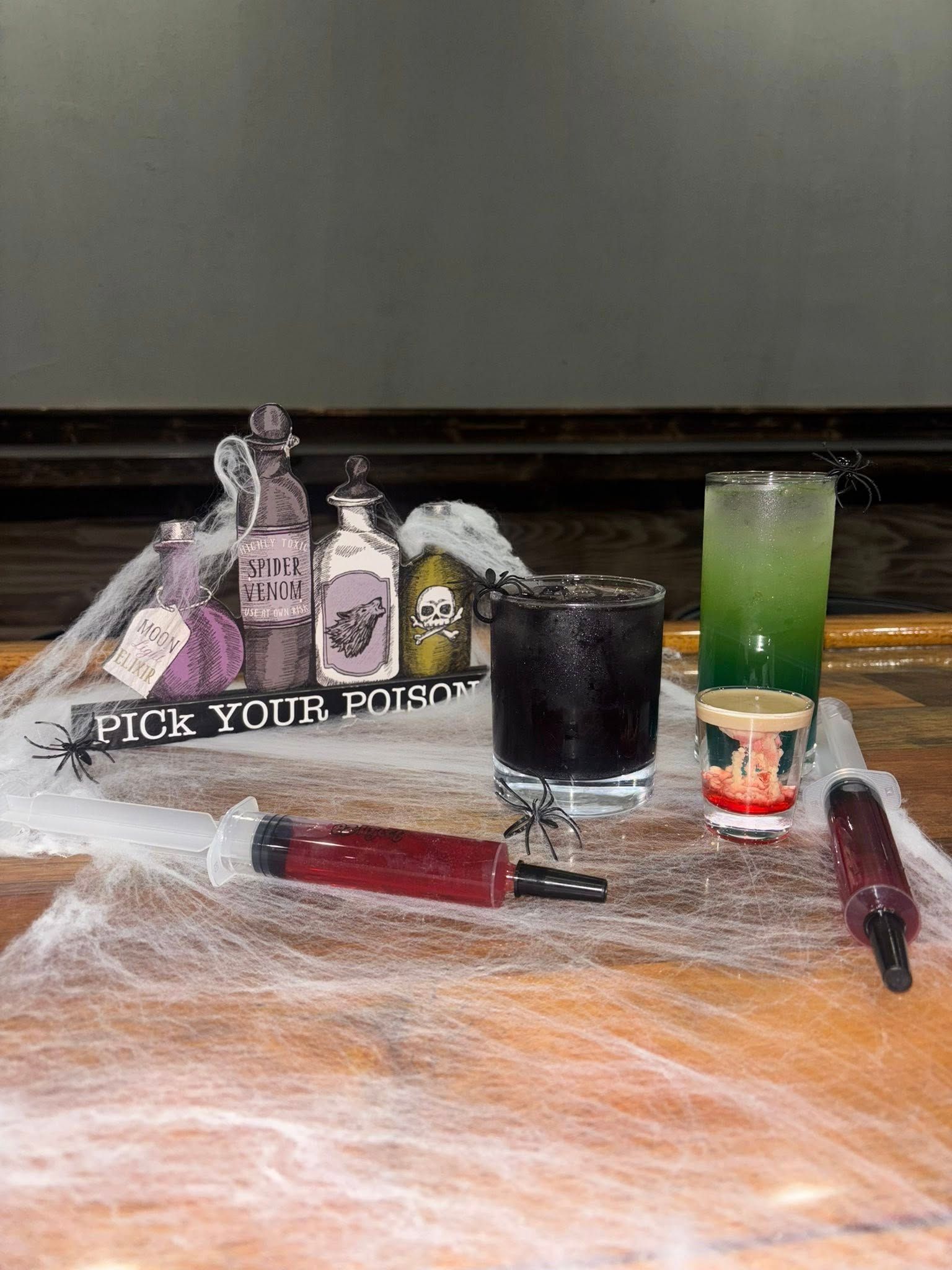 Halloween drinks display: Bottles, syringe, spider, and cocktails on a cobweb-covered table.