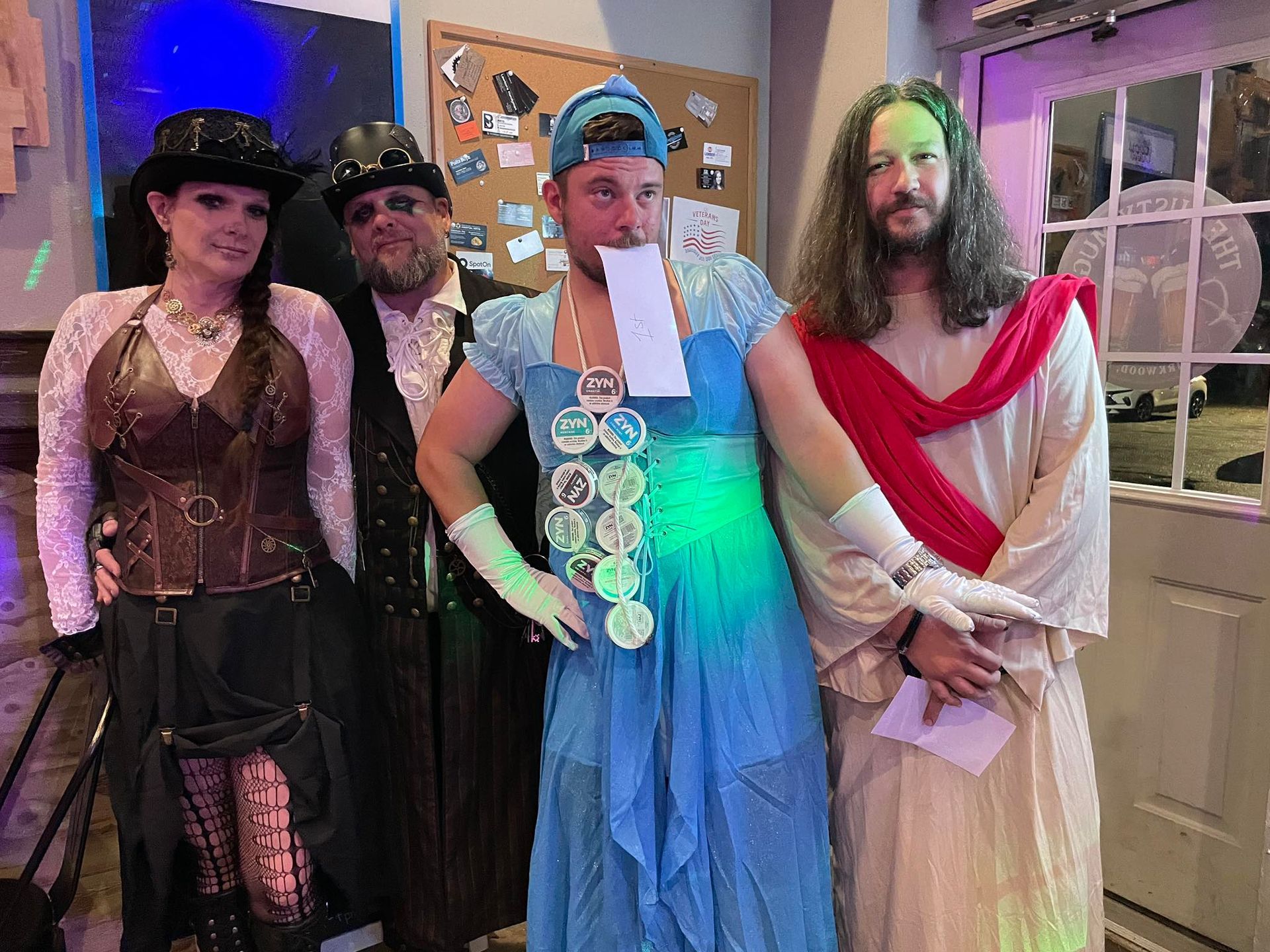Four people in costumes pose indoors. One in a steampunk outfit, one in a suit, one in a blue dress, and one dressed as Jesus.