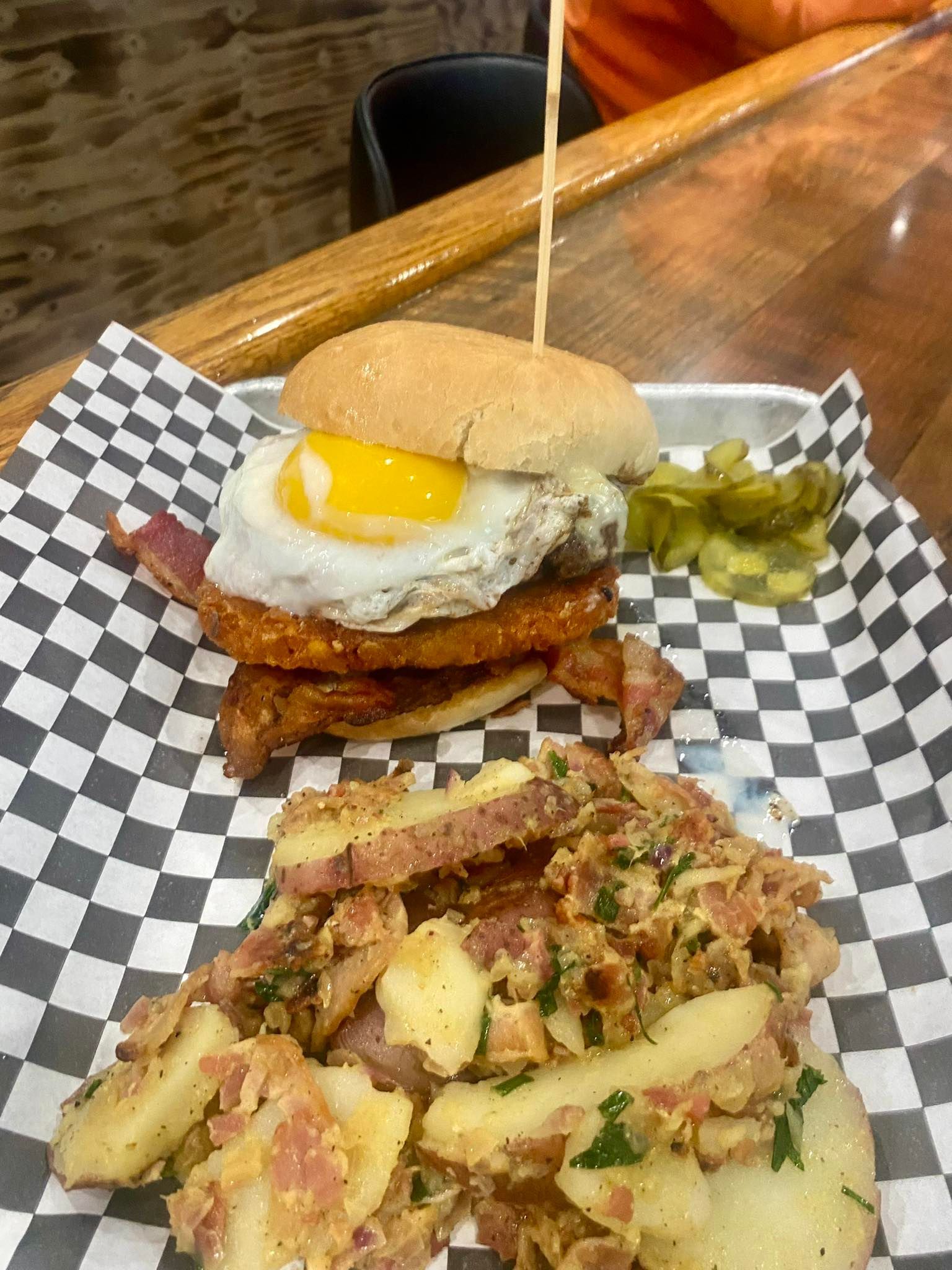 Burger with fried egg and bacon, potato salad, pickles on checkered paper.