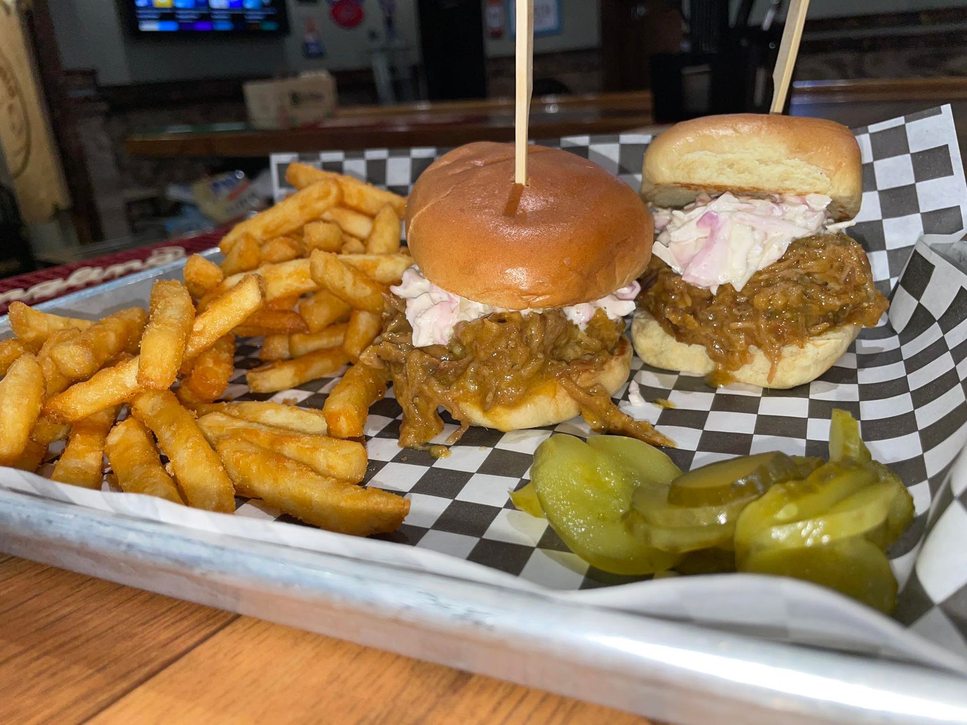 Pulled pork sliders with coleslaw on brioche buns, fries, and pickles on a checkered paper-lined tray.