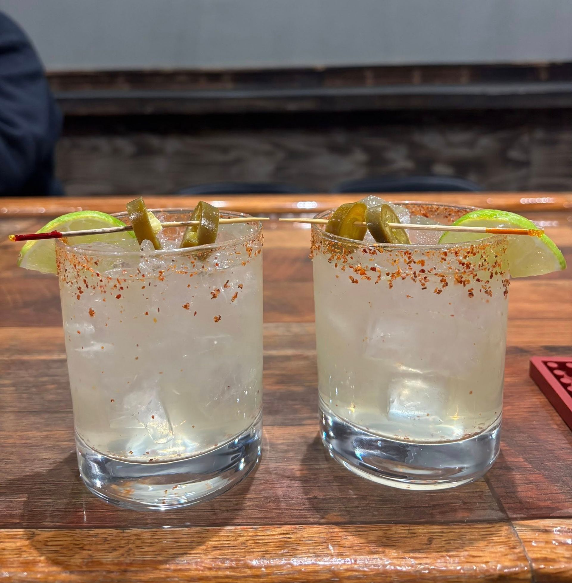 Two clear cocktails with lime and jalapeño garnishes on a wooden bar, rimmed with seasoning.