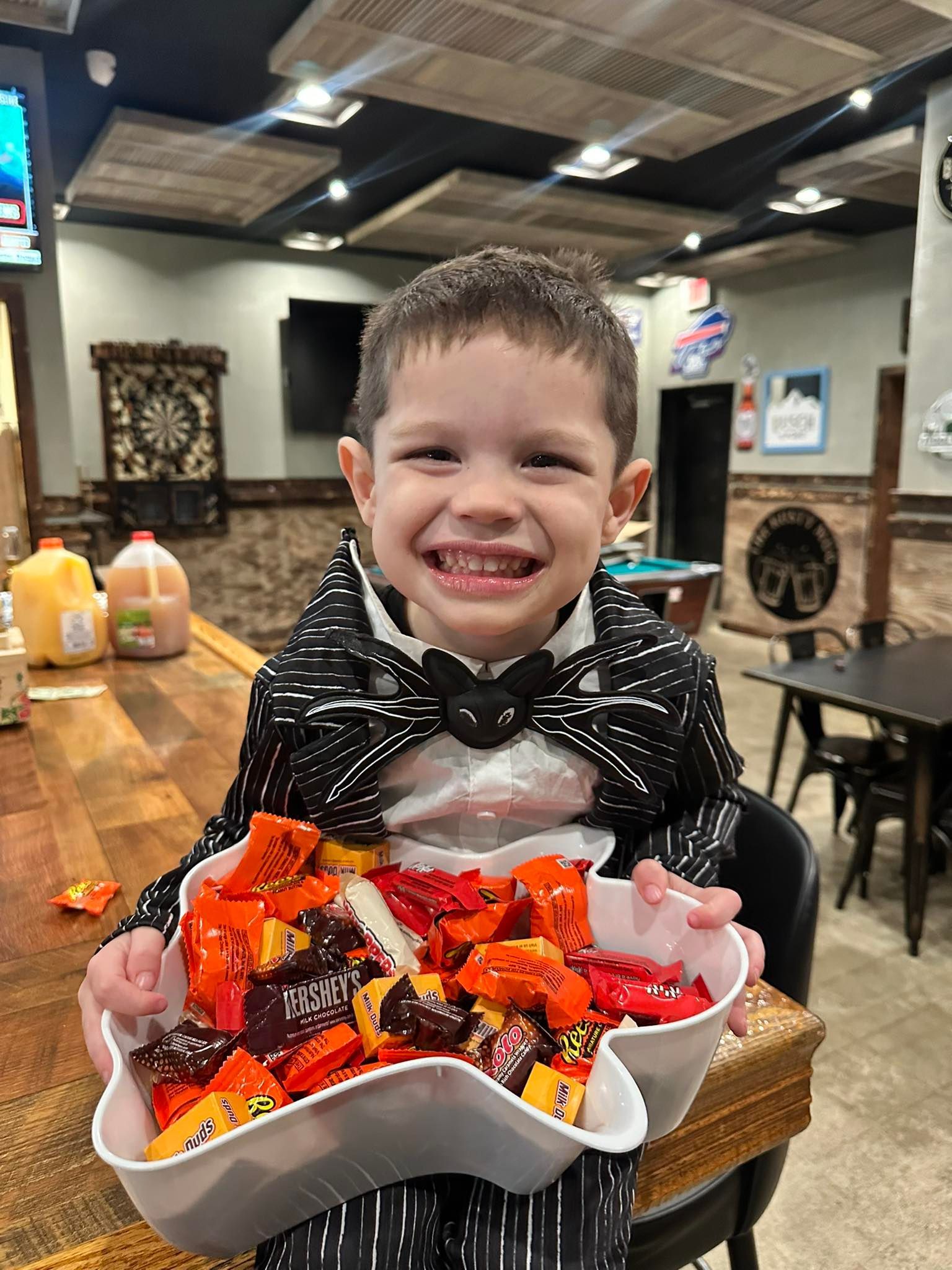Child in Halloween costume smiles, holding bowl of colorful candy. Interior, bar setting.