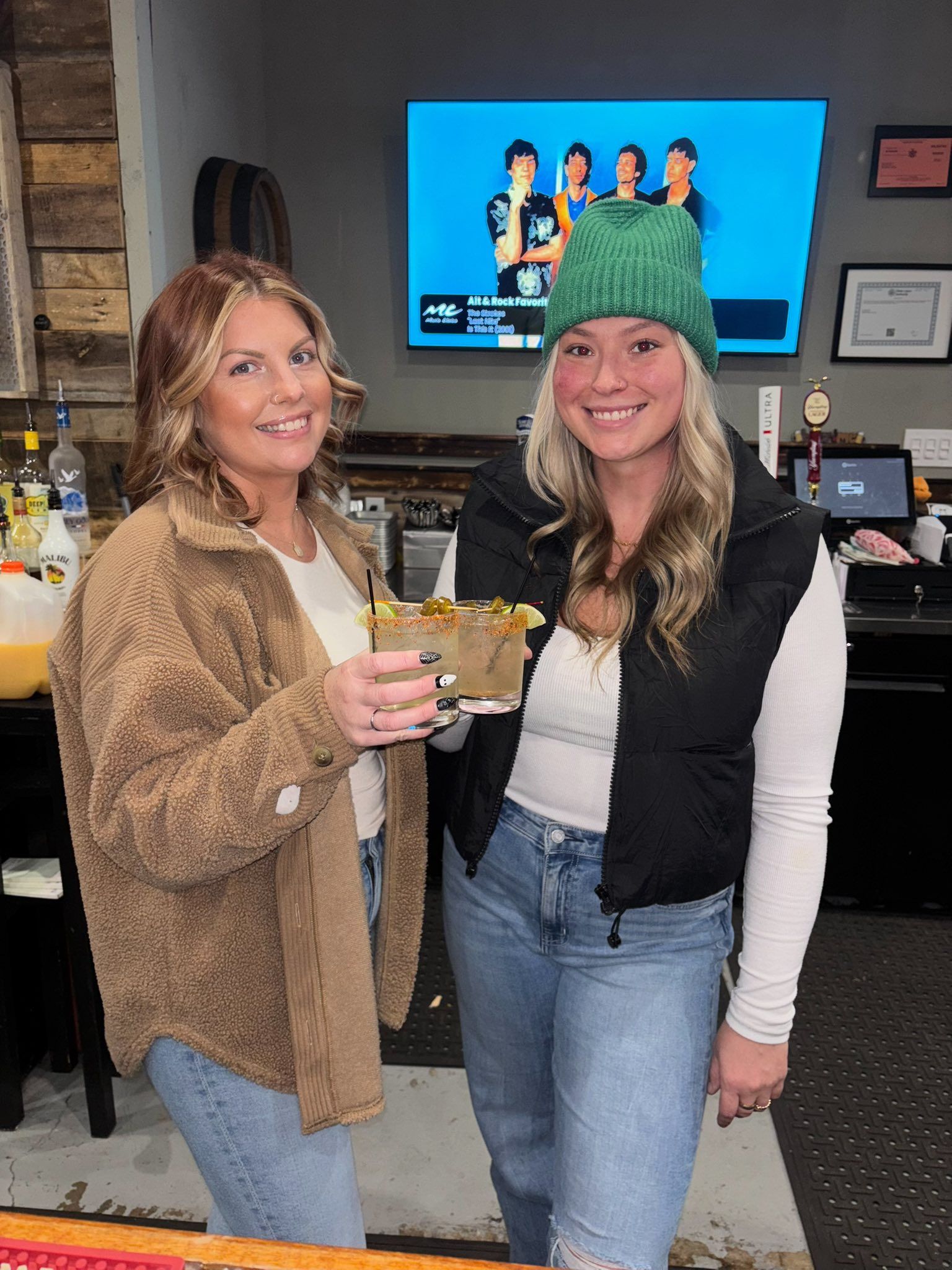 Two women in a bar hold a food tray; one wears a green hat. TV in the background.