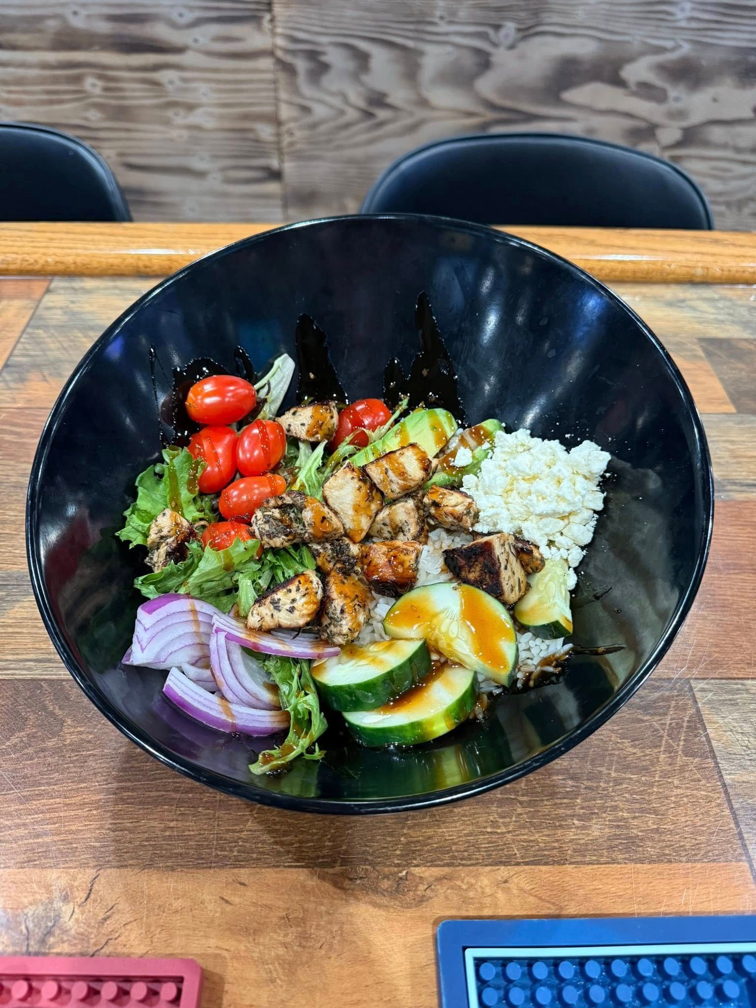 Chicken and vegetable salad bowl on a wooden table. Black bowl contains lettuce, tomatoes, cucumbers, onions, and grains.