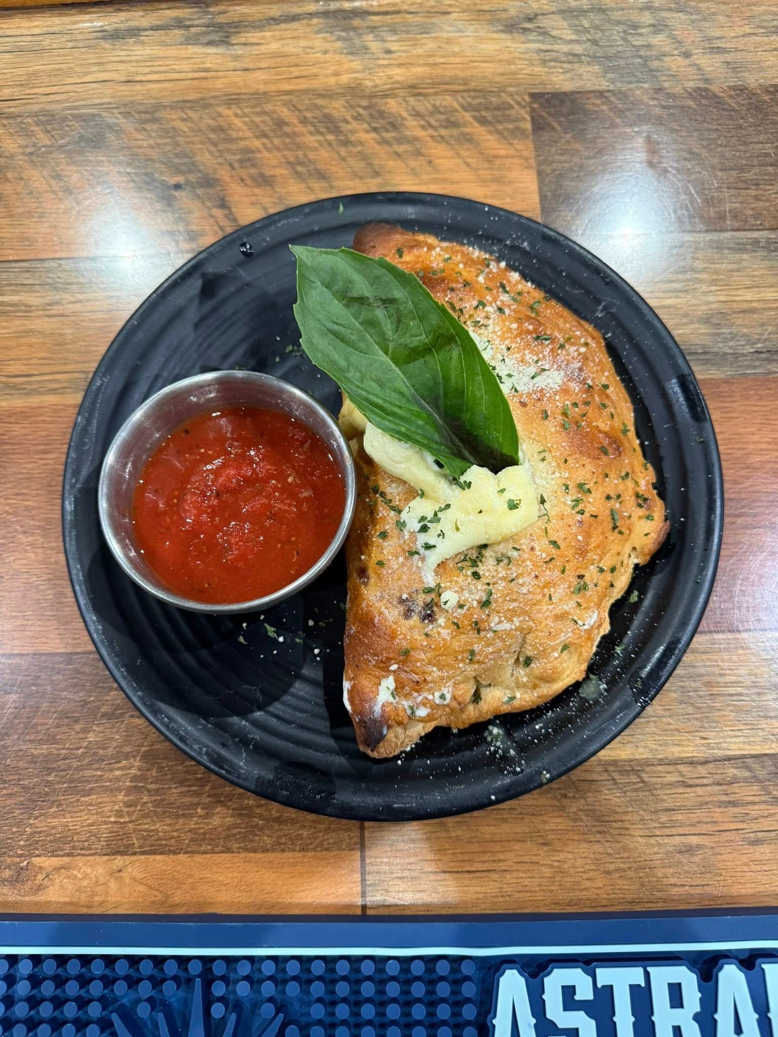 Calzone on a black plate with a small bowl of red sauce and basil garnish.