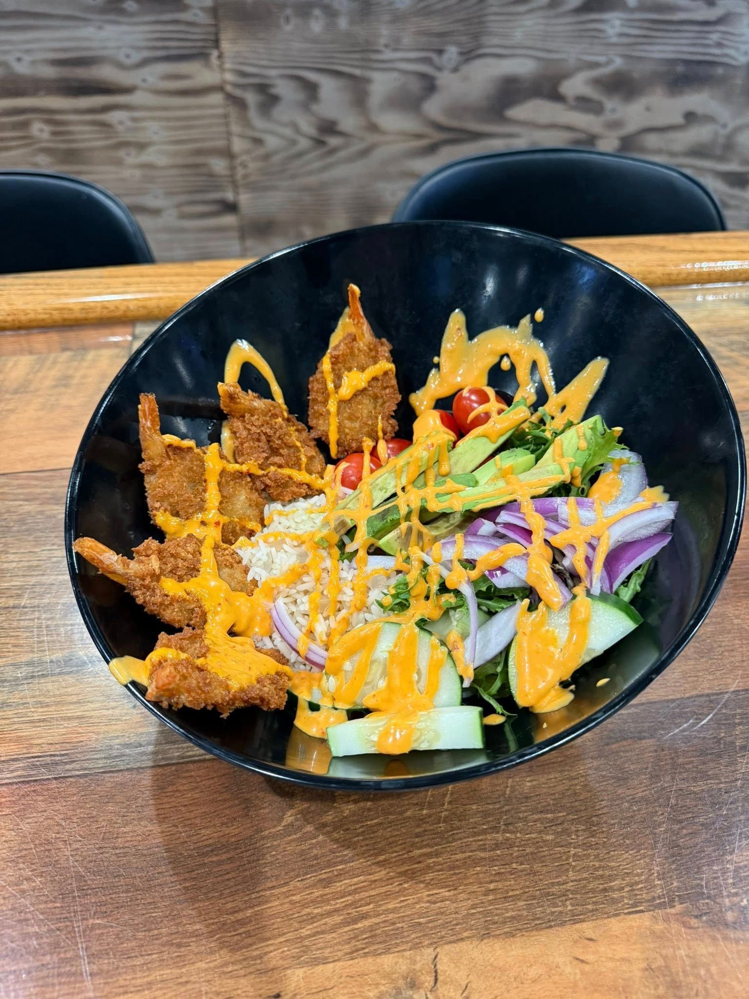 Black bowl with salad, fried shrimp, red onion, cucumbers, tomatoes, avocado, and orange drizzle.