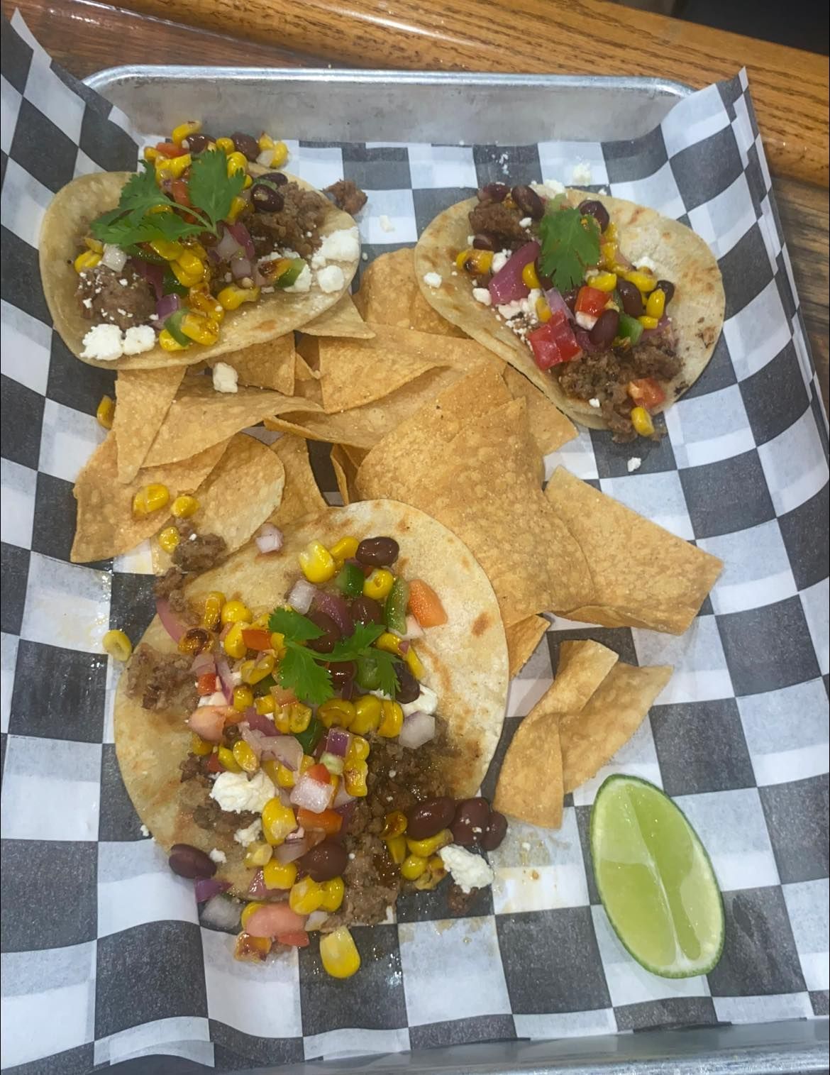 Tacos with toppings, chips, and lime wedge on checkered paper-lined tray.