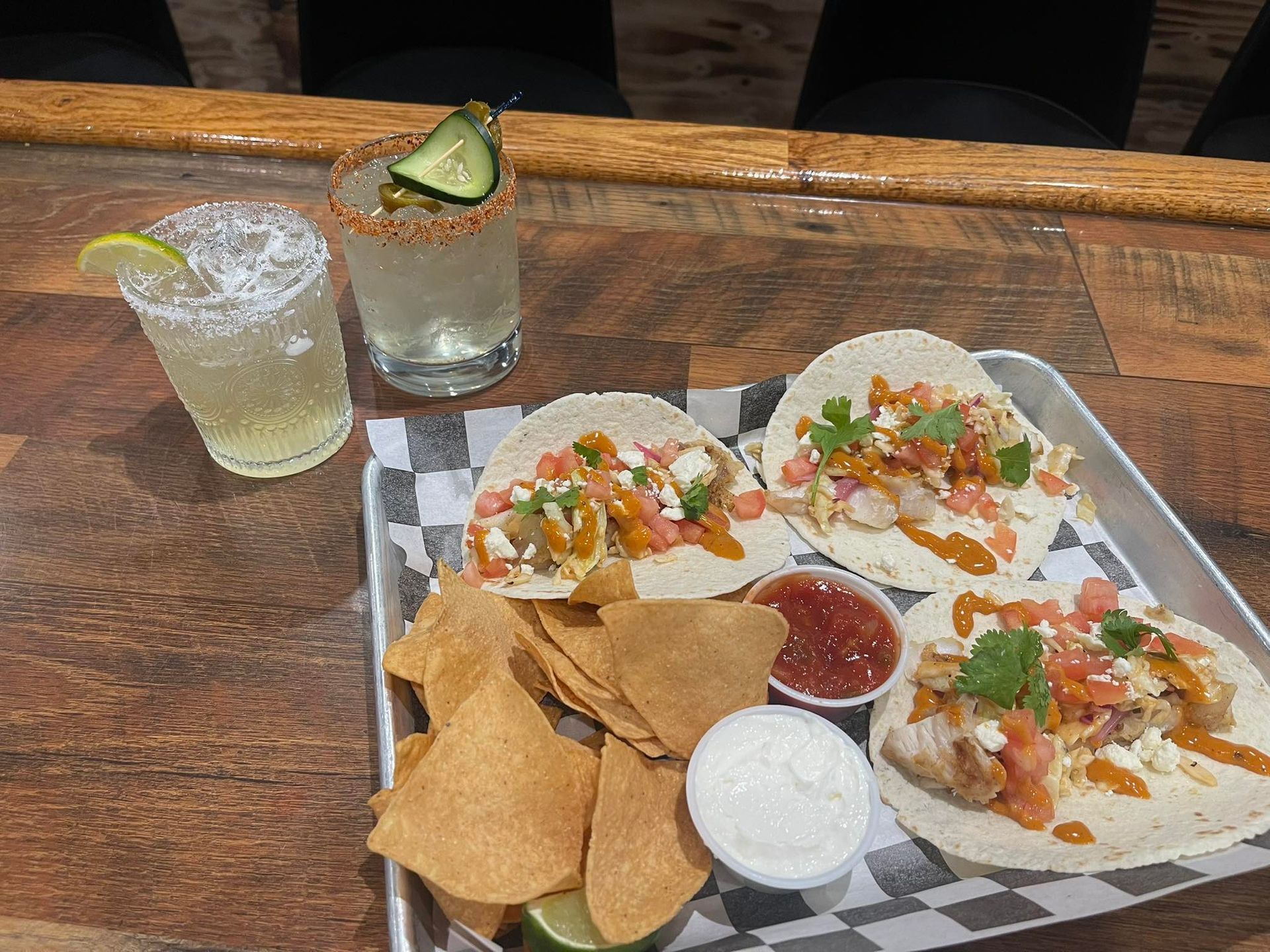 Margaritas, tacos, and tortilla chips on a bar top. Food tray includes salsa, sour cream, and lime wedges.