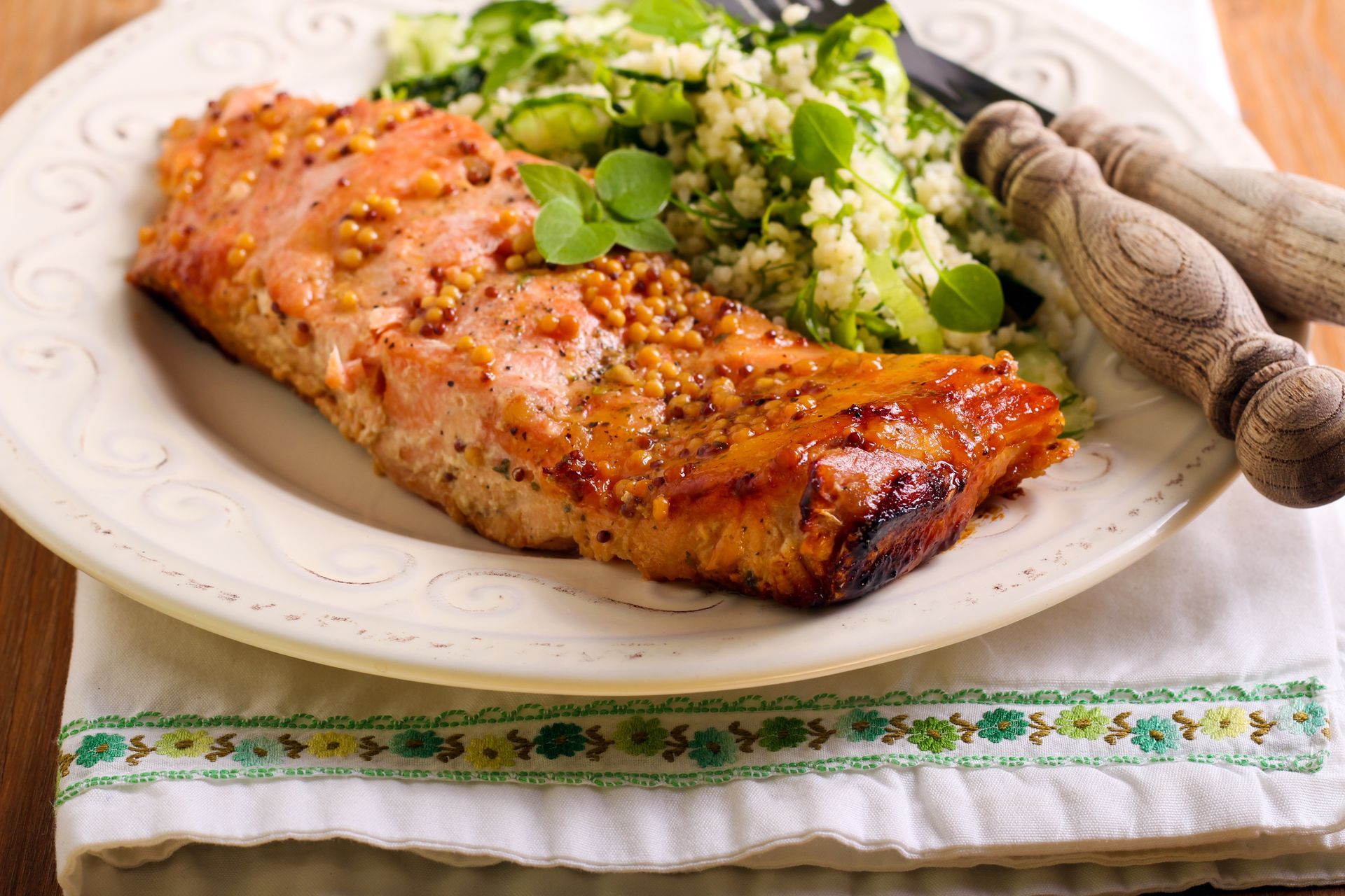 A cooked salmon fillet topped with mustard seeds served with a side of herbed couscous on a white plate.
