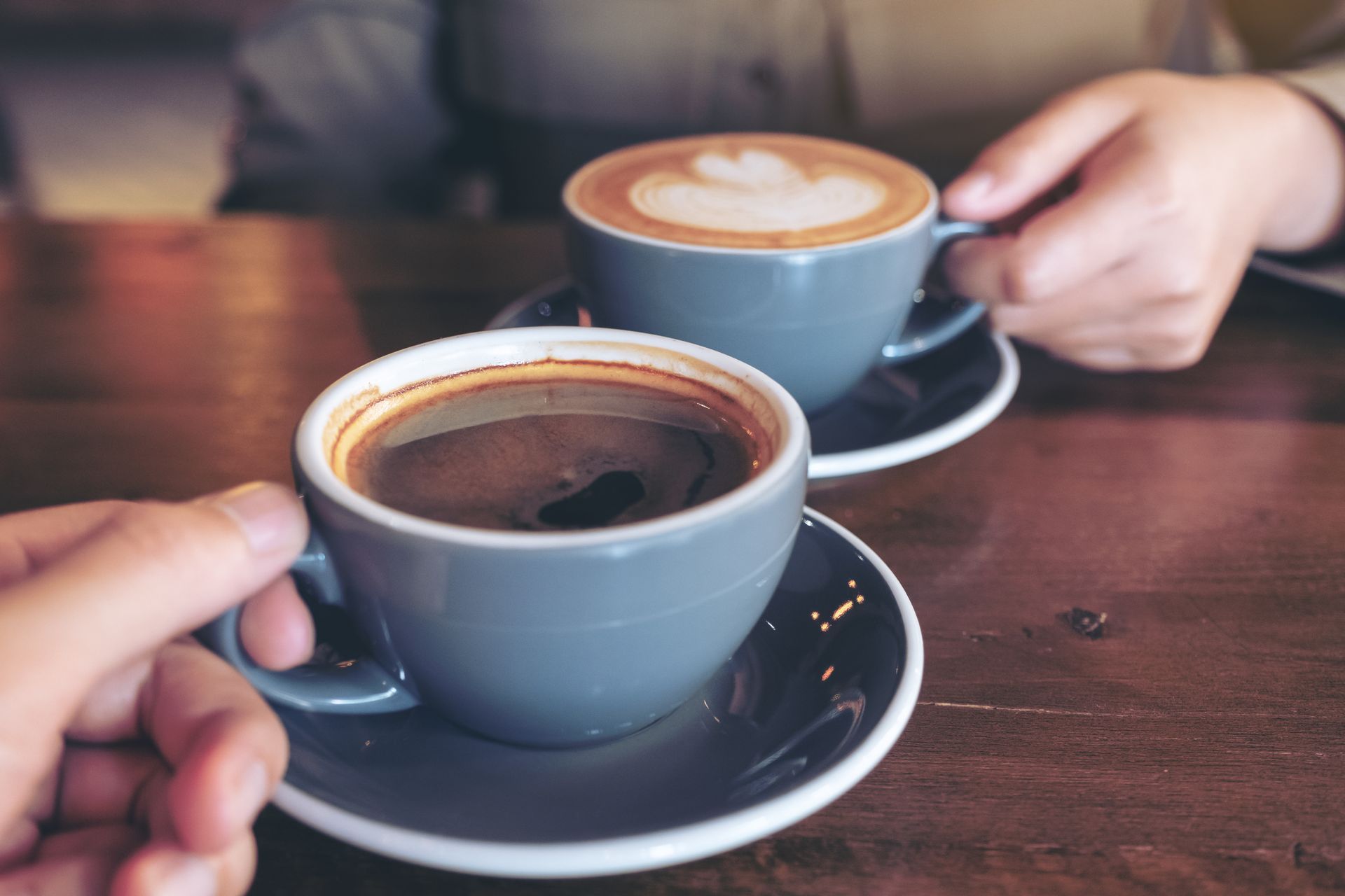 Two people hold gray ceramic cups of coffee on a wooden table, with one cup featuring latte art.