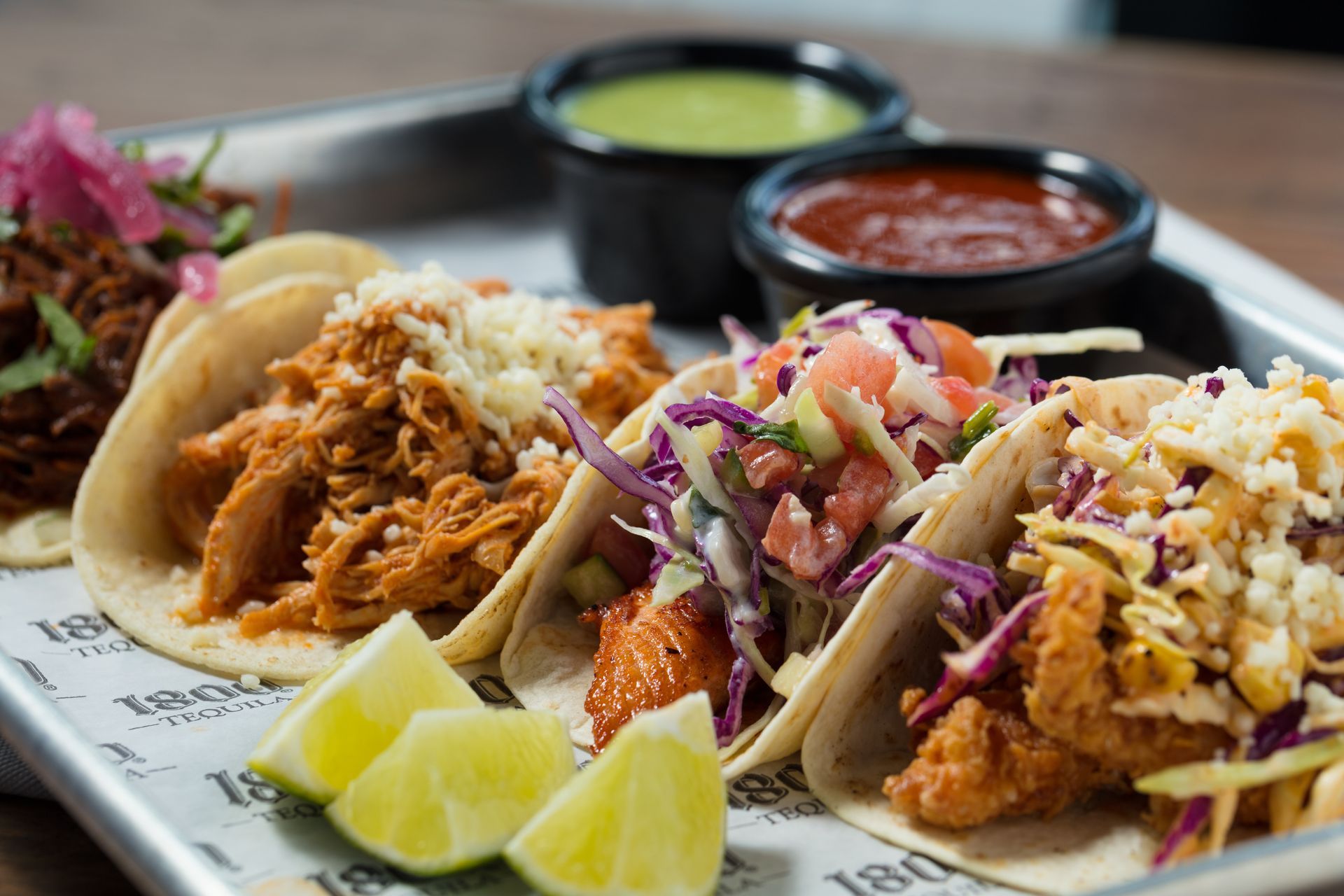 A metal tray features four diverse tacos with varied toppings, accompanied by two small bowls of green and red salsa.
