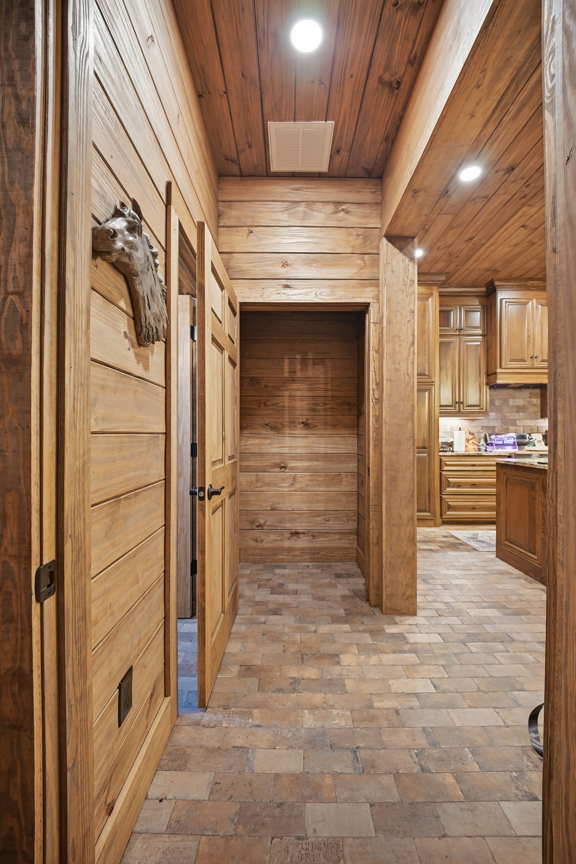 A hallway with wood-paneled walls and ceiling, stone tile flooring, and an open doorway leading into a kitchen.