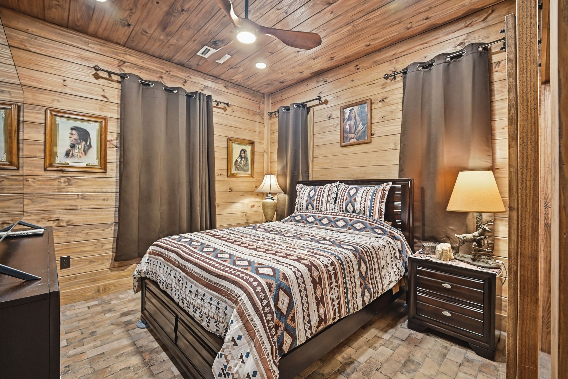 A rustic bedroom with wood-paneled walls, a bed featuring a patterned quilt, brown curtains, and a nightstand with a lamp.