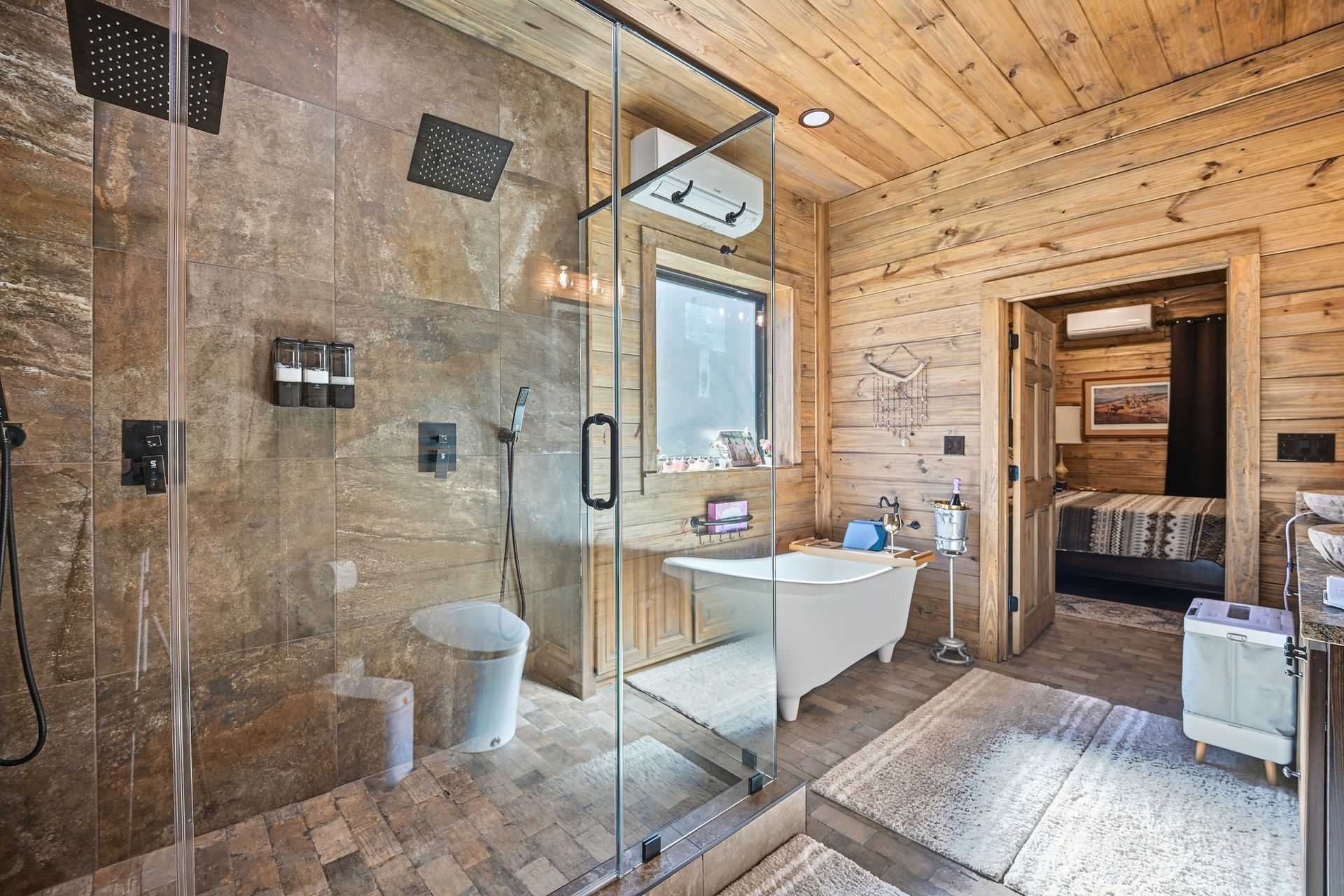 A rustic bathroom with a glass-enclosed shower, a white clawfoot bathtub, and light wood-paneled walls.