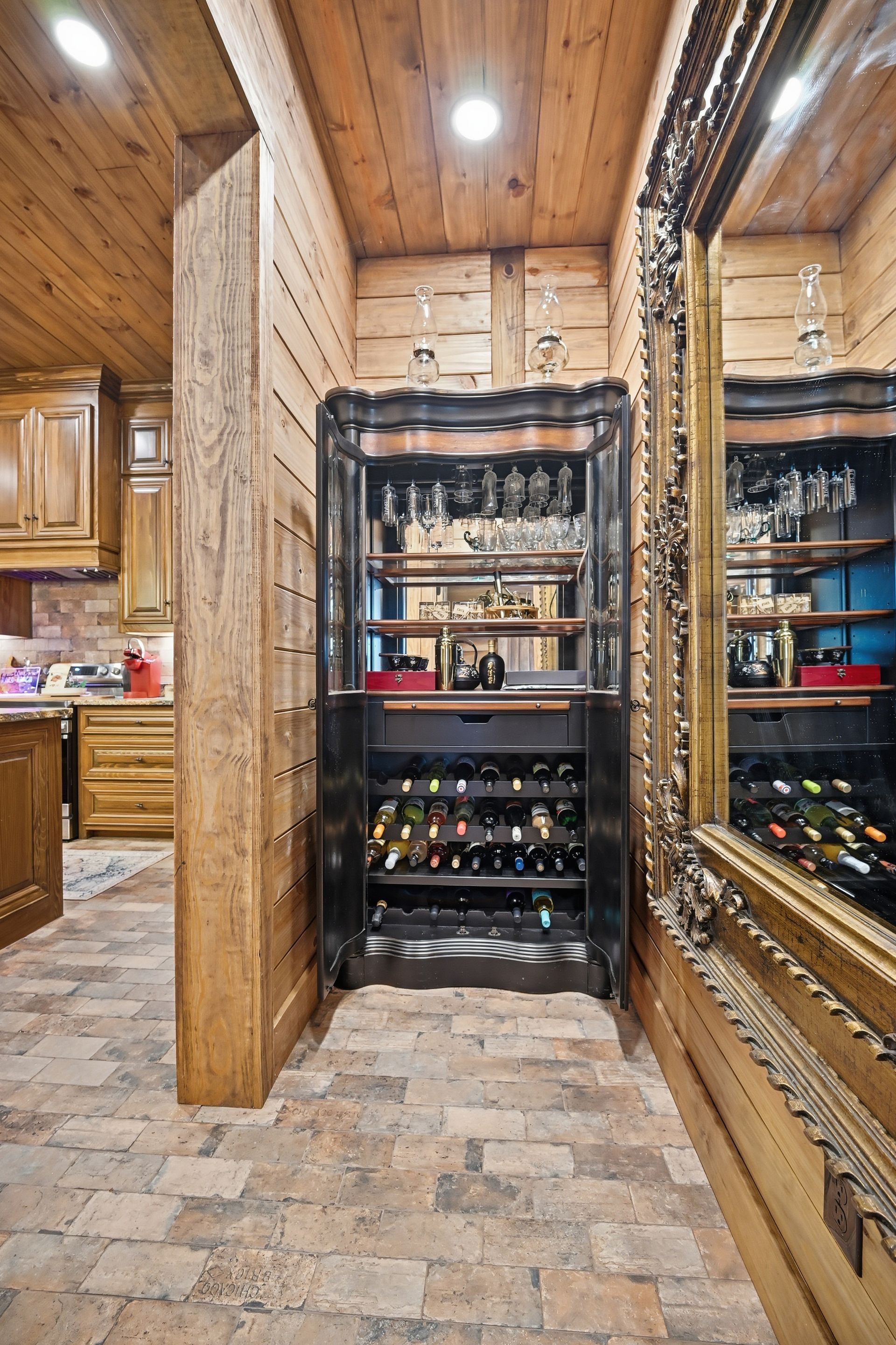A wine cabinet with glassware and bottles sits in a rustic room with wood-paneled walls and a brick floor.