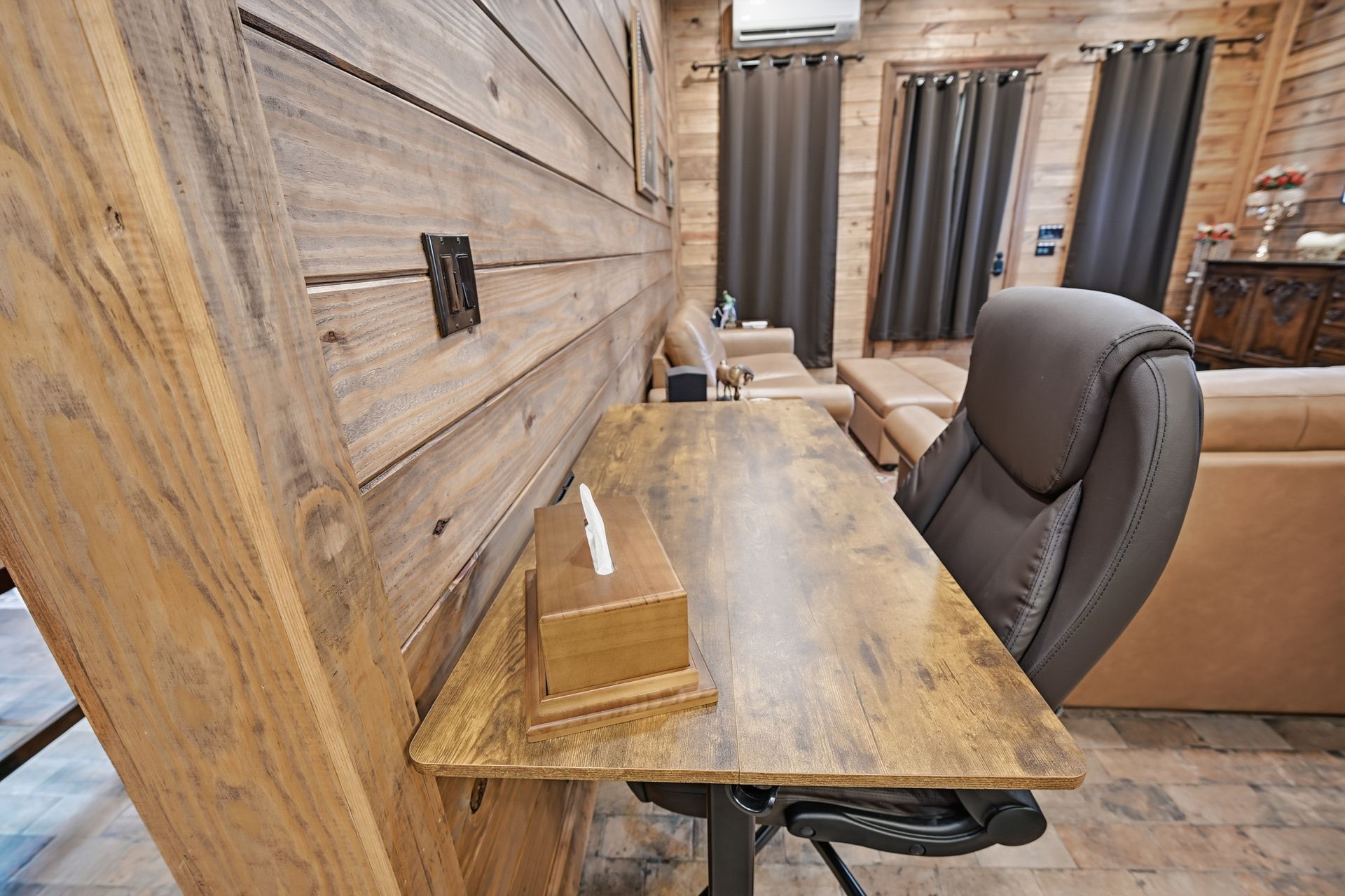 A wooden desk with a tissue box holder sits in front of a rustic, wood-paneled wall, accompanied by an office chair.
