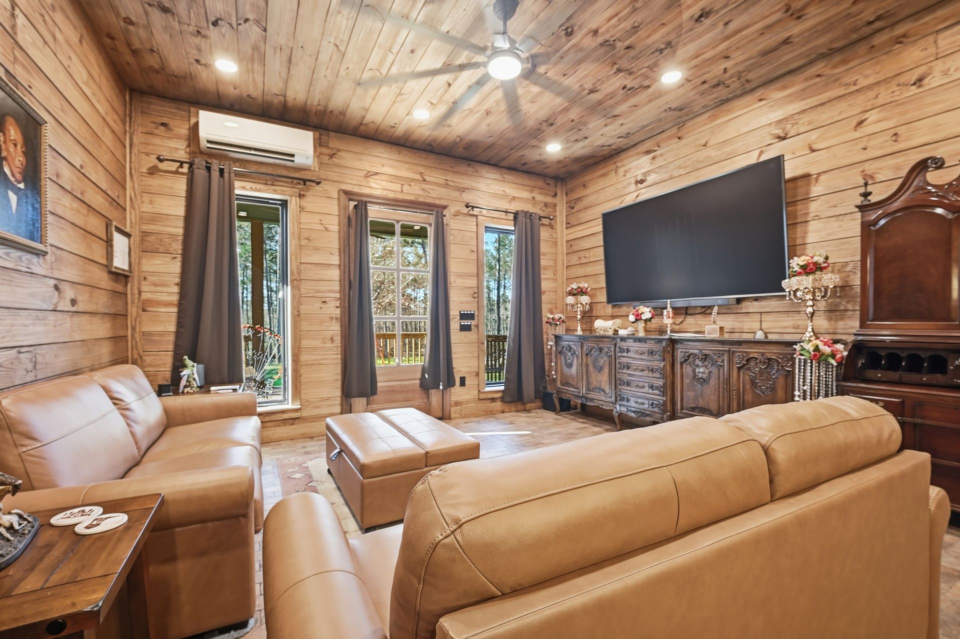 A rustic living room with log walls, two tan leather sofas, a matching ottoman, a large wall-mounted TV, and a ceiling fan.