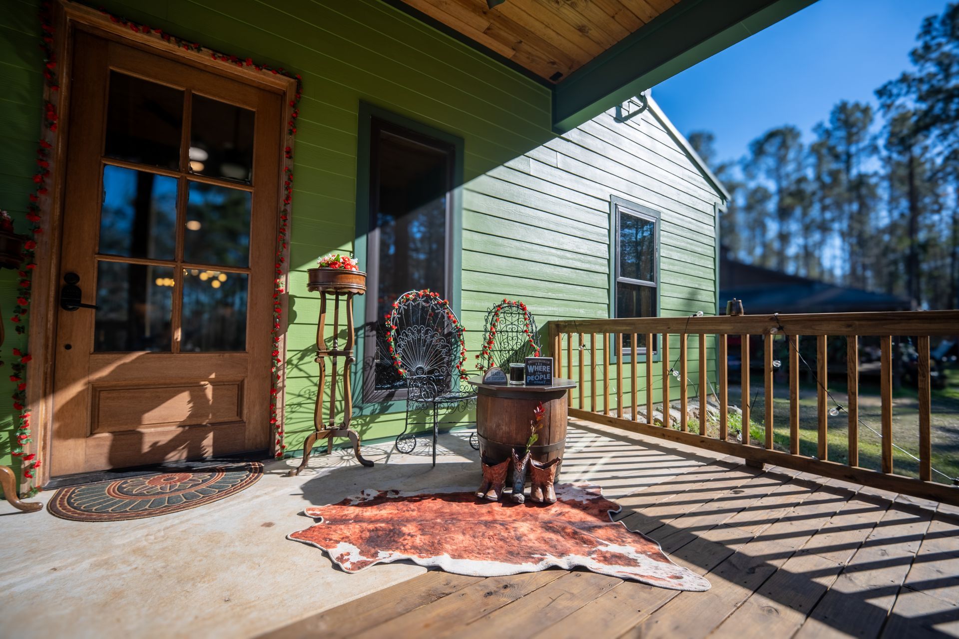 A green wooden porch features a wooden door, a metal chair, a decorative side table, and a cowhide rug on the floor.