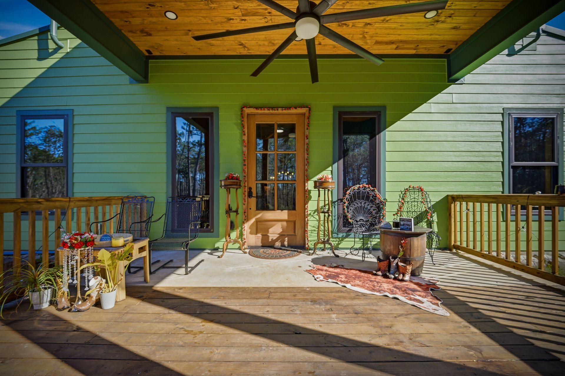 A porch with green wooden walls, a wood-paneled ceiling with a fan, outdoor furniture, and a central wooden front door.