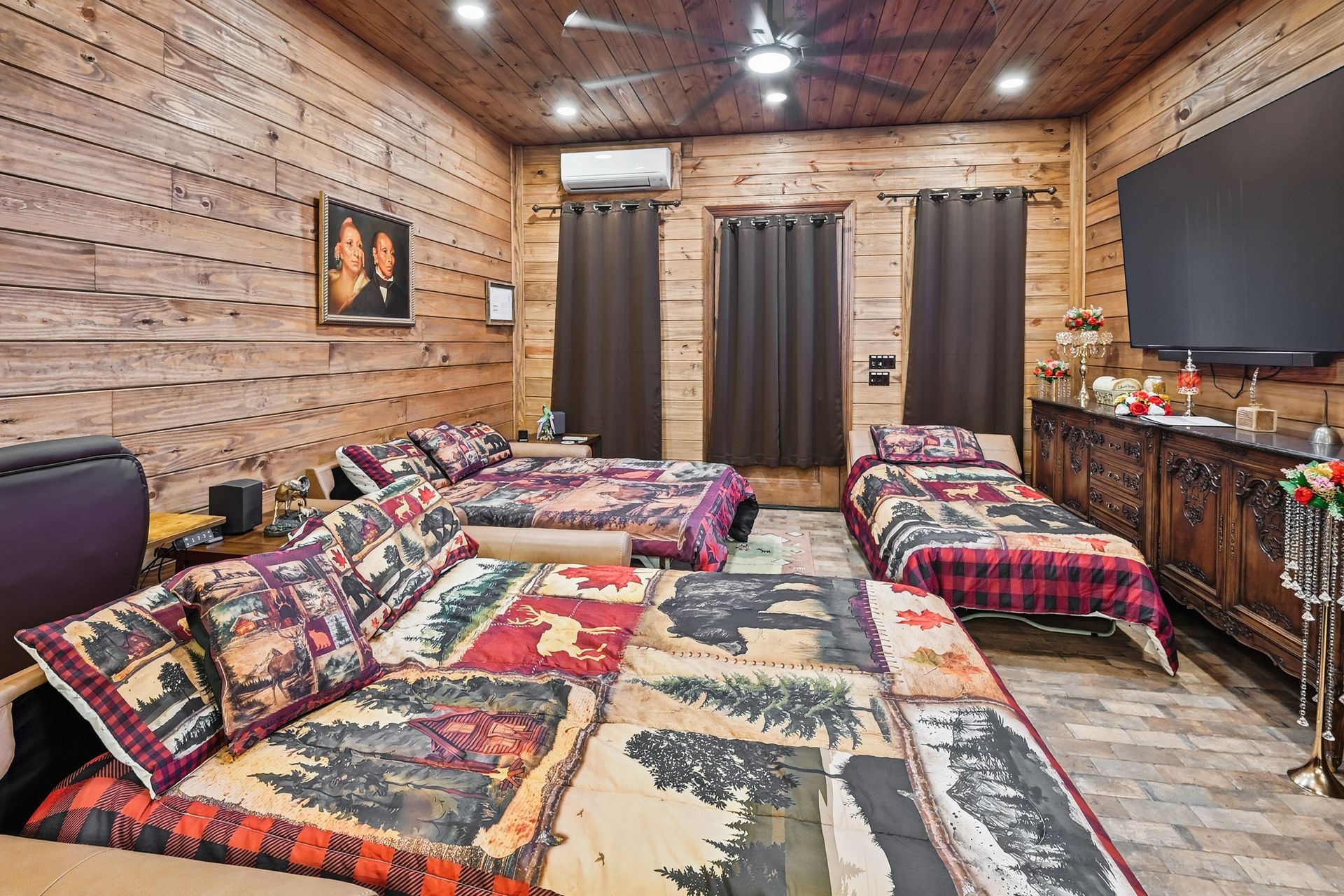 A rustic bedroom features wood-paneled walls, two beds with bear-themed quilts, dark curtains, and a large wall-mounted TV.