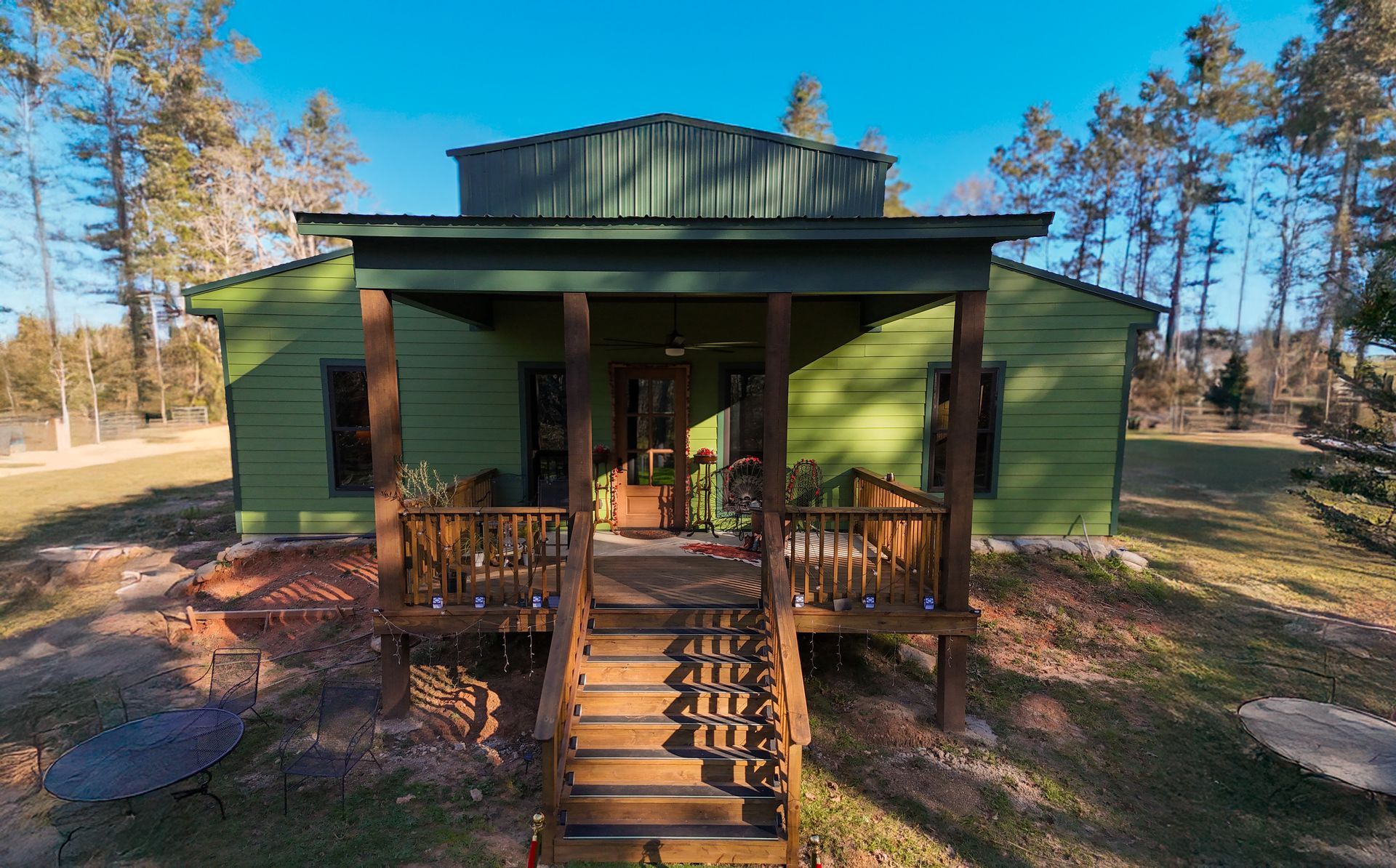 A sage-green cabin with a raised wooden porch and stairs, surrounded by a sunny, tree-filled landscape.
