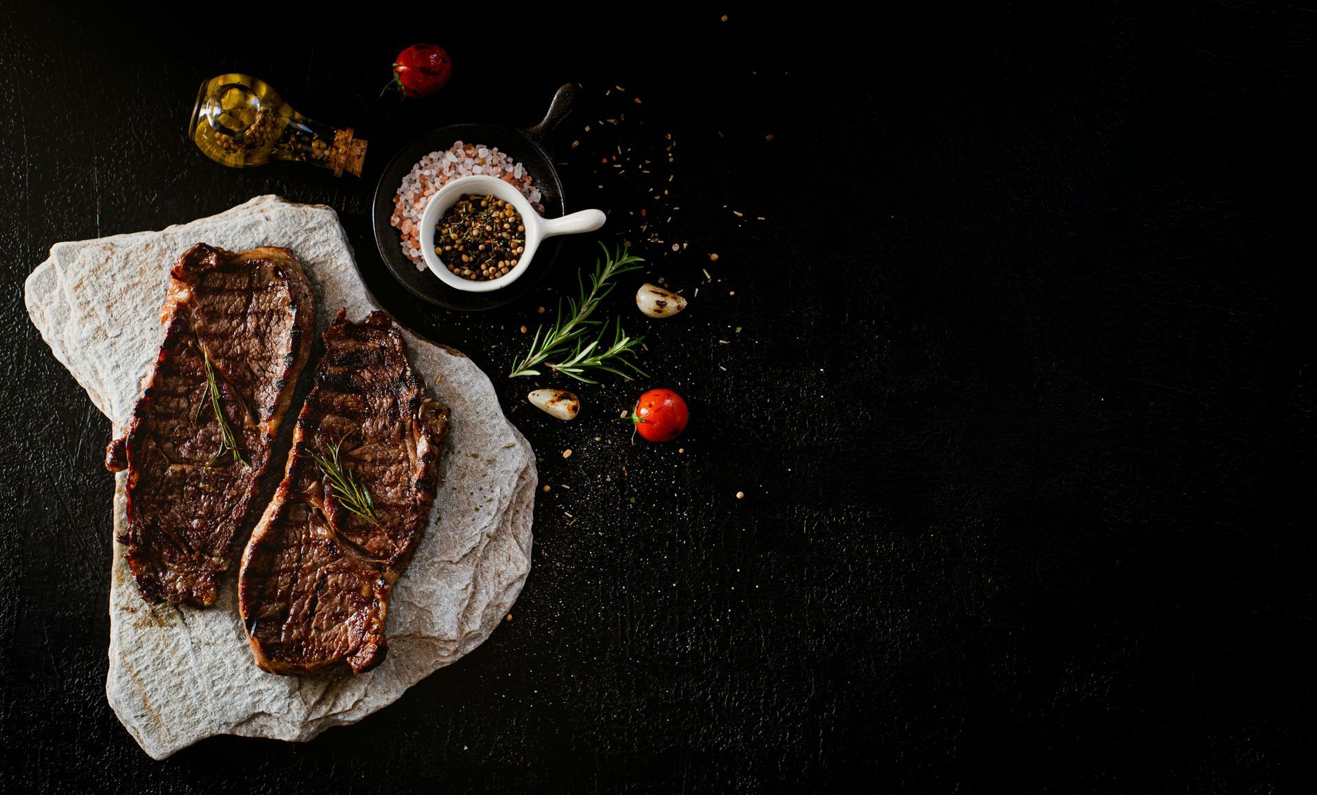 Two grilled steaks on a stone slab with seasoning, herbs, and oil against a black background.