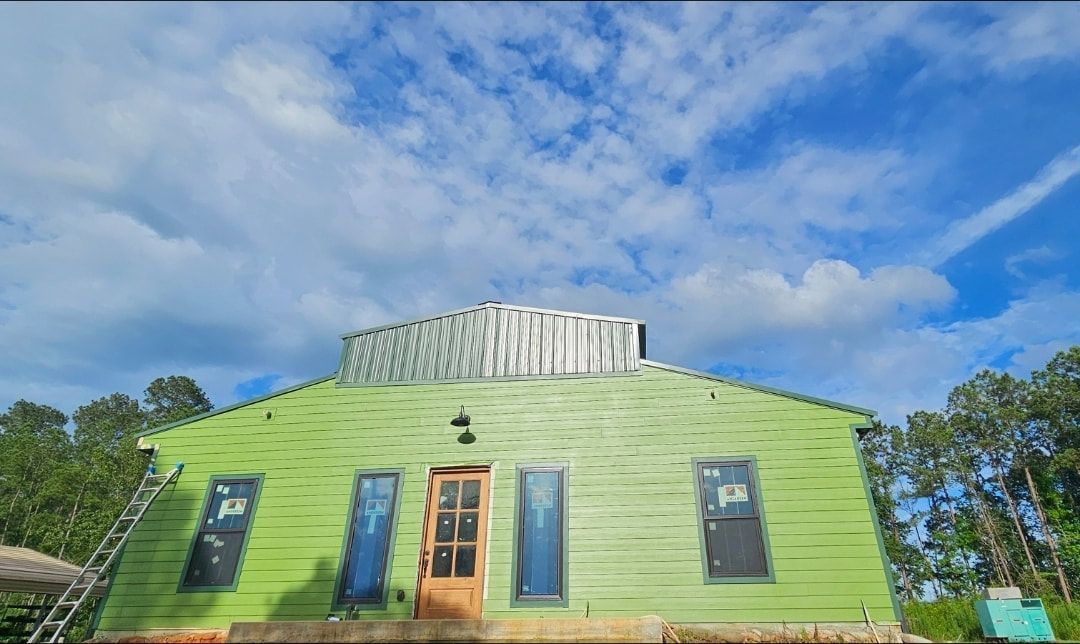 A light green, wooden building with a wooden door and four windows against a bright, cloudy blue sky.
