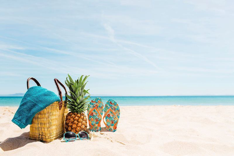 Beach tote, pineapple, sandals, and colorful flip-flops on a white sandy beach by turquoise water