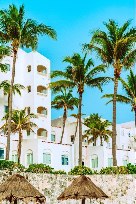 White resort buildings with palm trees and thatched umbrellas by a sunny beach under a blue sky