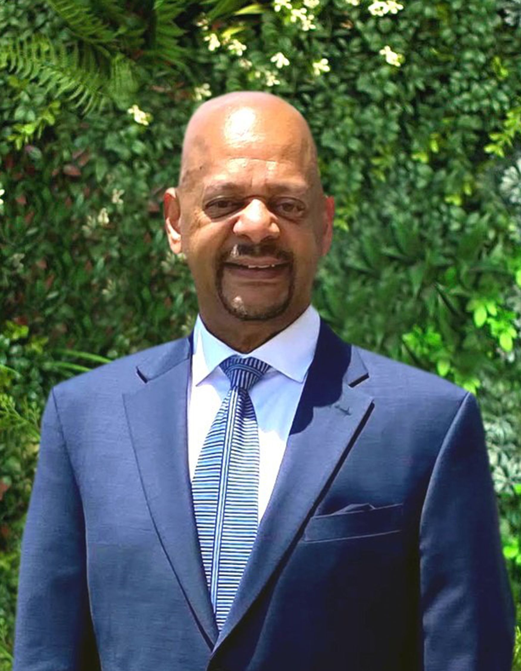 A man in a suit and tie is standing in front of a wall of plants.