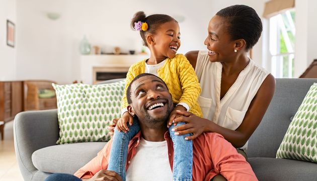 A man and woman are sitting on a couch with a little girl on his shoulders.