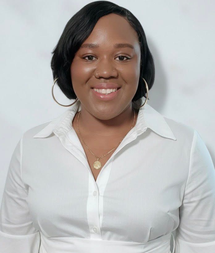 A woman wearing a white shirt and a necklace is smiling in front of a green wall.