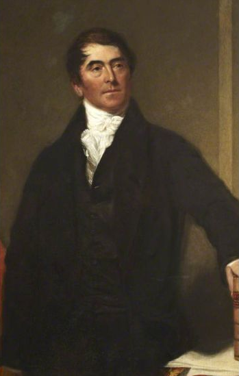 Portrait of George Birbeck in dark clothing, white cravat. Leaning on a desk, neutral background.