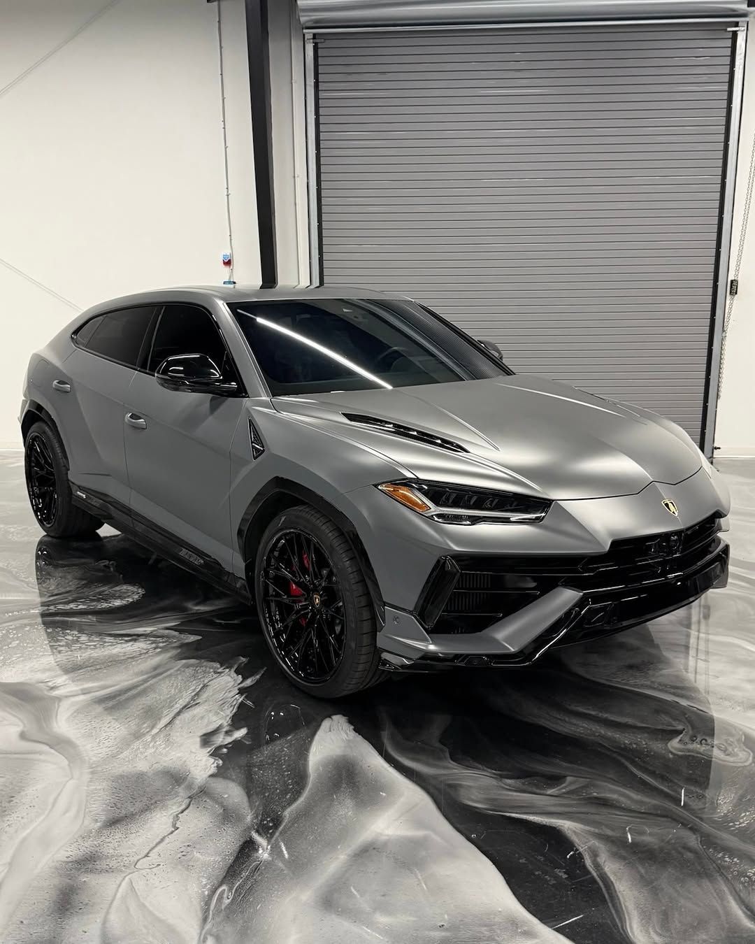 Gray Lamborghini Urus SUV parked in a garage with a reflective epoxy floor and a closed gray roller door.