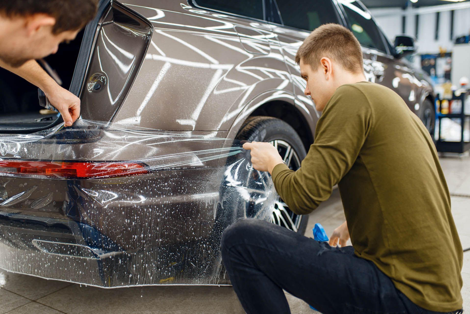 Two people applying protective film to a brown car's bumper in a shop.
