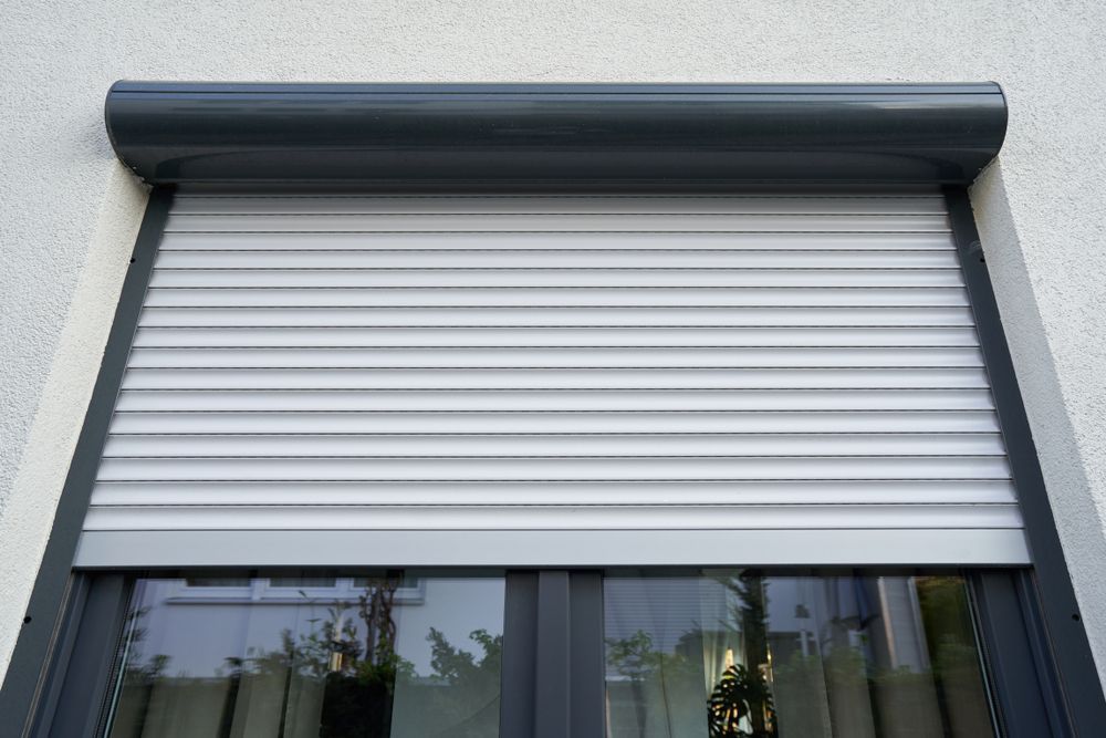 A gray roller shutter partially covers a window on a light-colored building exterior  — LeSands Screens & Blinds Moss Vale Pty Ltd in Moss Vale, NSW