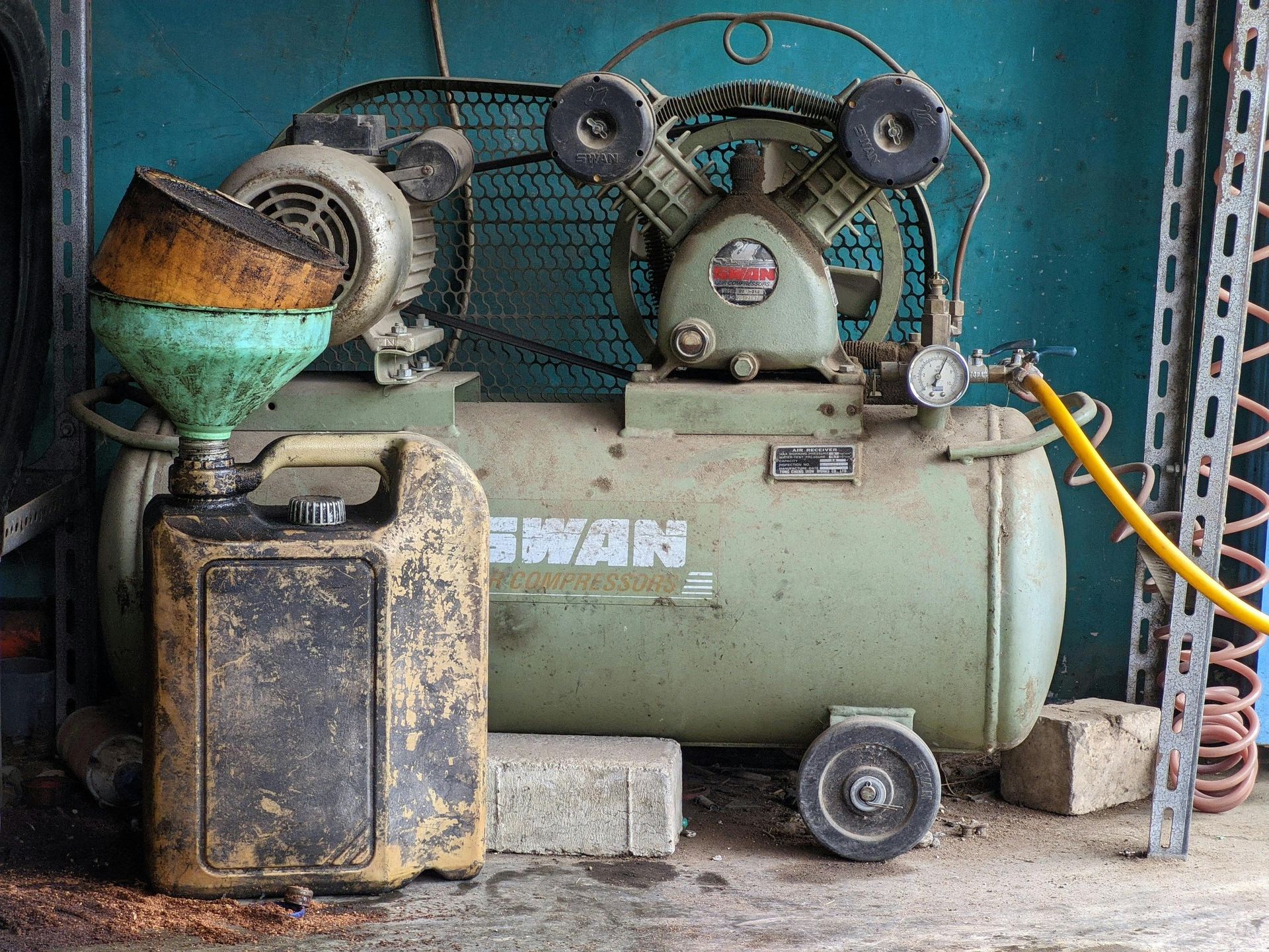 Dusty air compressor and machinery in a garage, with hoses, a wheelbarrow, and a fuel can.