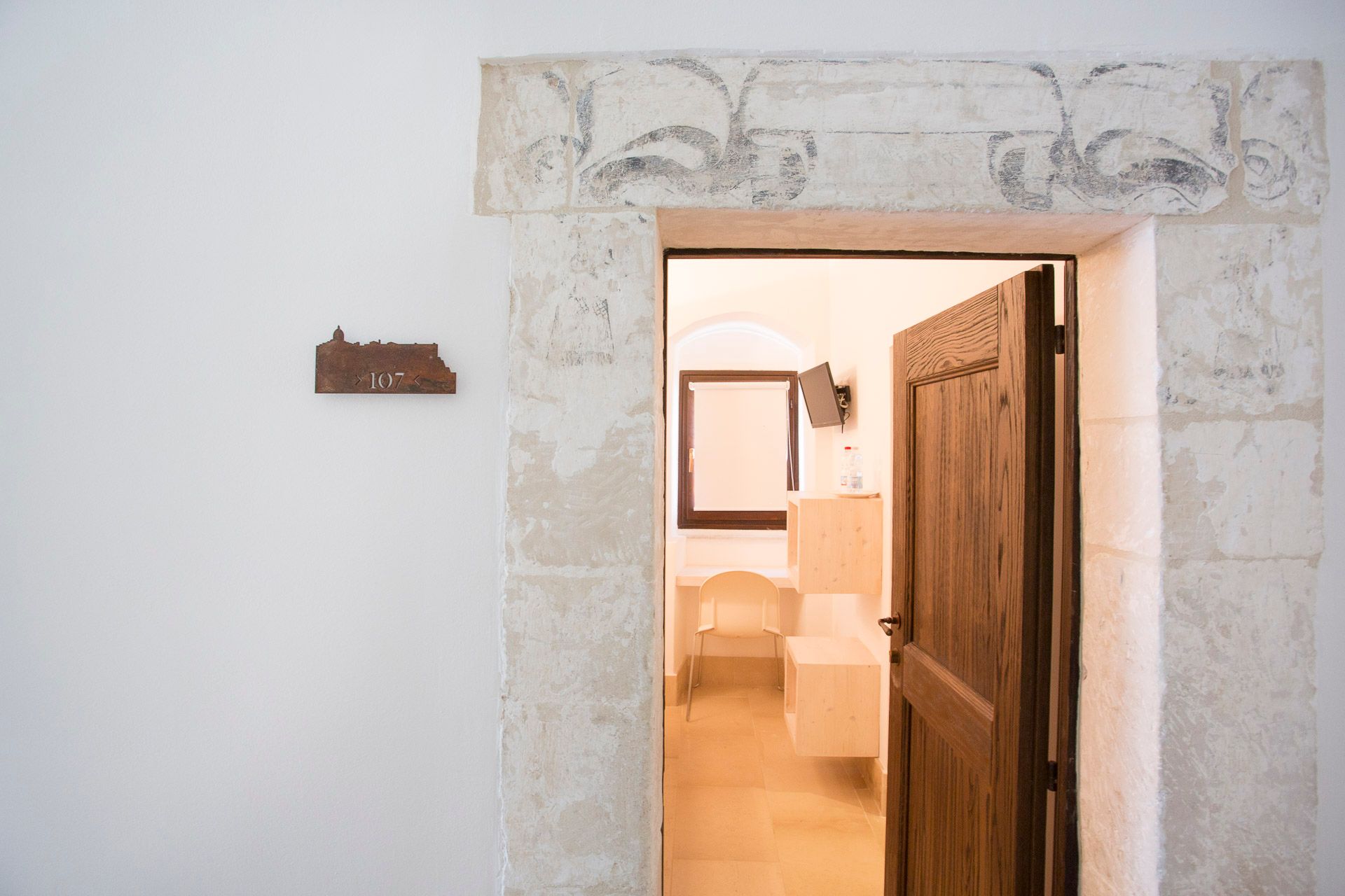 Una porta di legno è aperta su una stanza con un muro bianco. L'austerità delle stanze dei cappuccini | Antico Convento Ibla