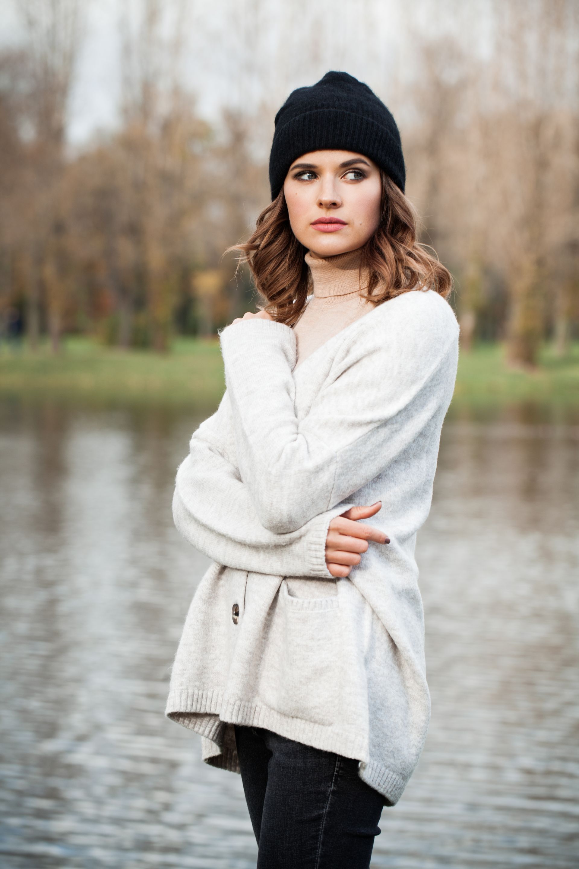 Una donna con un maglione bianco e un cappello nero è in piedi davanti a un lago.