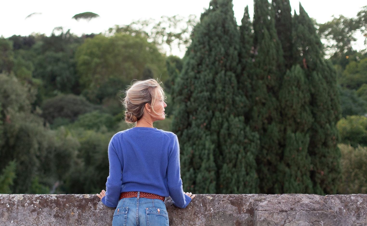 Una donna con un maglione blu e jeans è appoggiata a un muro.